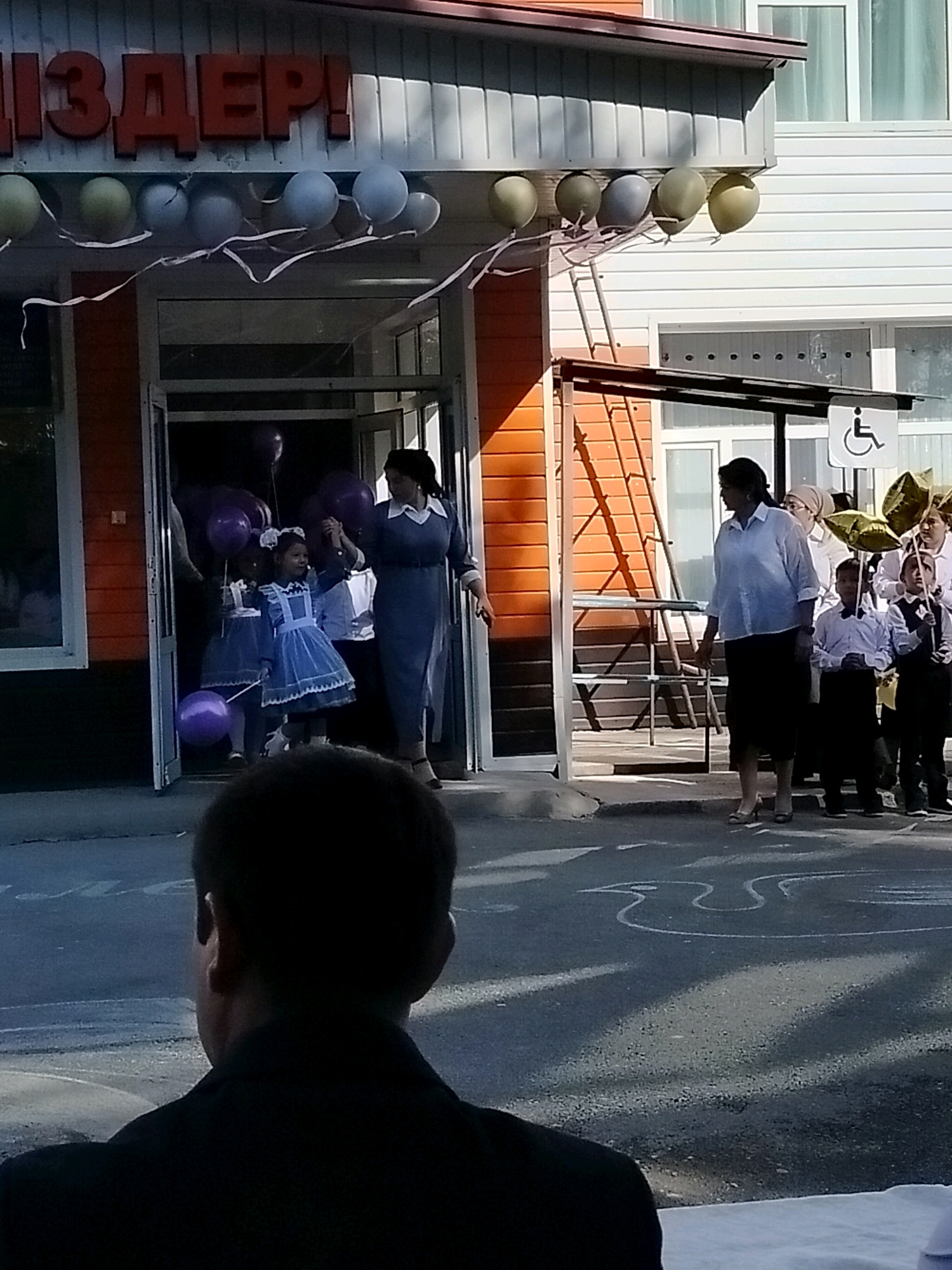 This image depicts an outdoor scene in front of a building with orange siding and a dark-colored awning. Red, three-dimensional lettering spelling "ЛИЗДЕР!" is mounted above the entrance. Several balloons are strung across the awning, some are olive green and others are light blue.
At the entrance, a woman in a dark blue dress with a white collar is holding the hand of a young girl wearing a light blue dress with a white apron and white hair accessories. The girl is holding a small purple balloon on a string. Behind them, other children and adults are visible, some holding balloons.
To the right of the entrance, a group of children in white shirts and dark pants or skirts are standing. One child holds a bunch of gold-colored balloons. A woman in a white shirt and dark skirt is standing nearby. A sign with a wheelchair accessibility symbol is visible on a pole.
The ground in front of the building is paved, and white chalk drawings, possibly of a bird and other shapes, are visible on the asphalt. In the foreground, the back of a person's head and shoulders, wearing a dark jacket, is visible. The overall setting appears to be a public gathering or event.