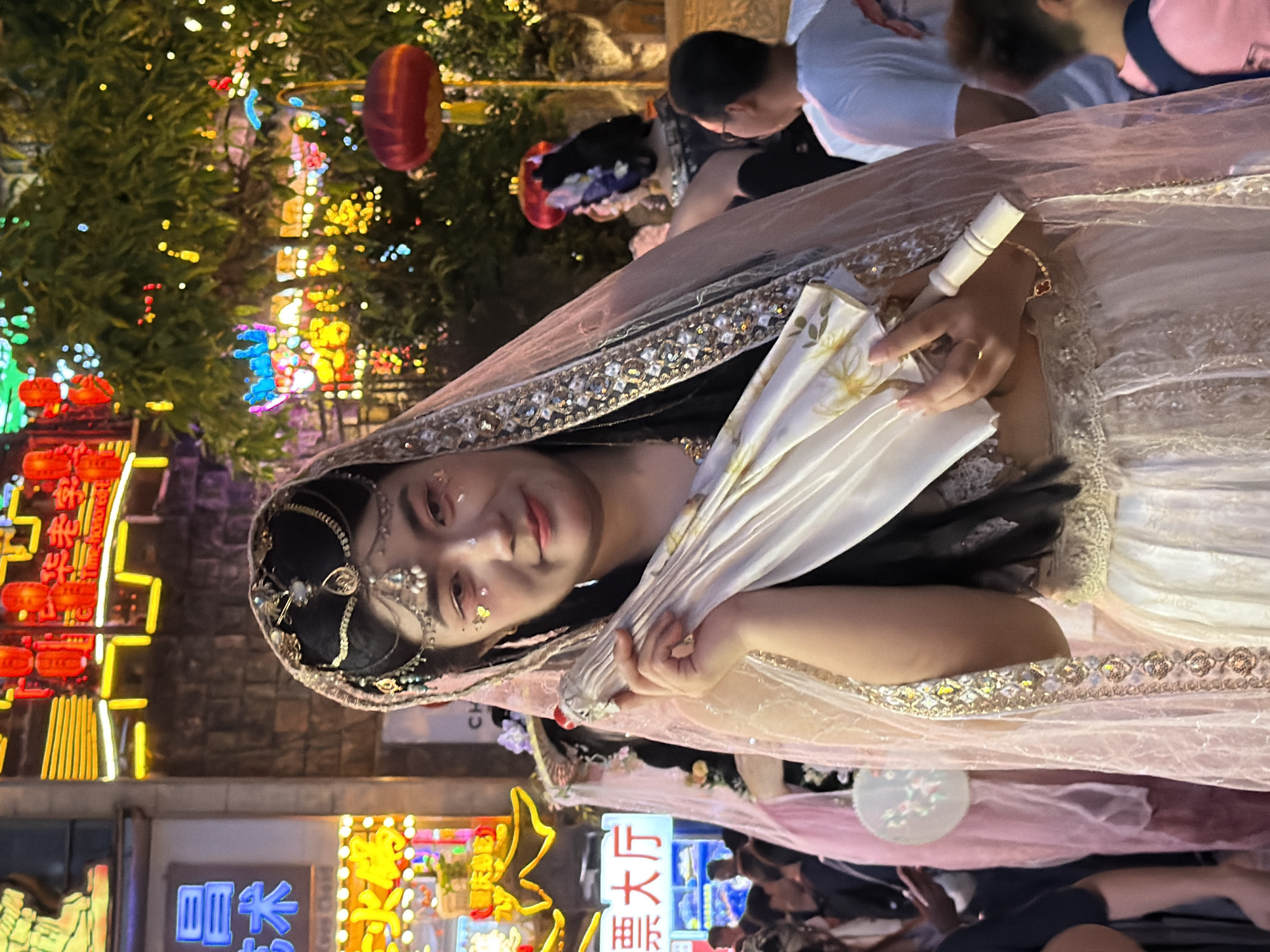 The image depicts a woman in traditional Indian attire, possibly a bride or a performer, adorned with jewelry and a veil. She is holding a folded white fan with a floral pattern. The background suggests a festive evening setting, likely in a market or street, with numerous colorful lights and lanterns illuminating the scene. Chinese characters are visible on illuminated signs, indicating a location in China, specifically Yuzhong District. The overall atmosphere appears lively and celebratory. The woman's facial expression is gentle and engaging, and the intricate details of her costume, including gold embellishments and delicate embroidery, are prominent. The lighting suggests it is nighttime.