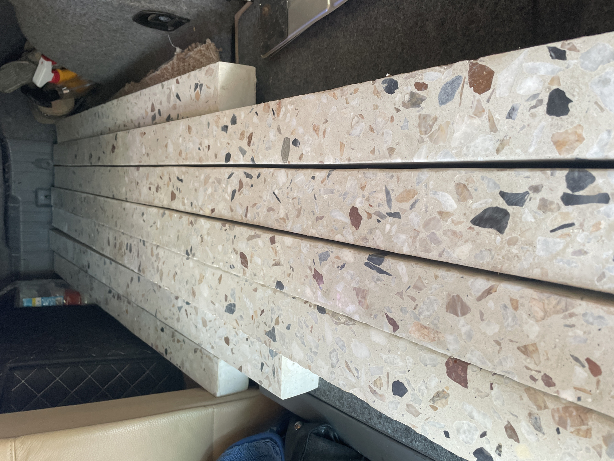 The image shows a stack of four rectangular, light-colored terrazzo-like slabs loaded into the back of a vehicle. The slabs are resting on white foam spacers. The background consists of the interior of a vehicle, including the upholstered side panel and some items obscured in shadow. The vehicle's cargo area is lined with grey fabric. It is not possible to ascertain a specific location within Krasnodar, Russia, or any information about people involved, based on the image.