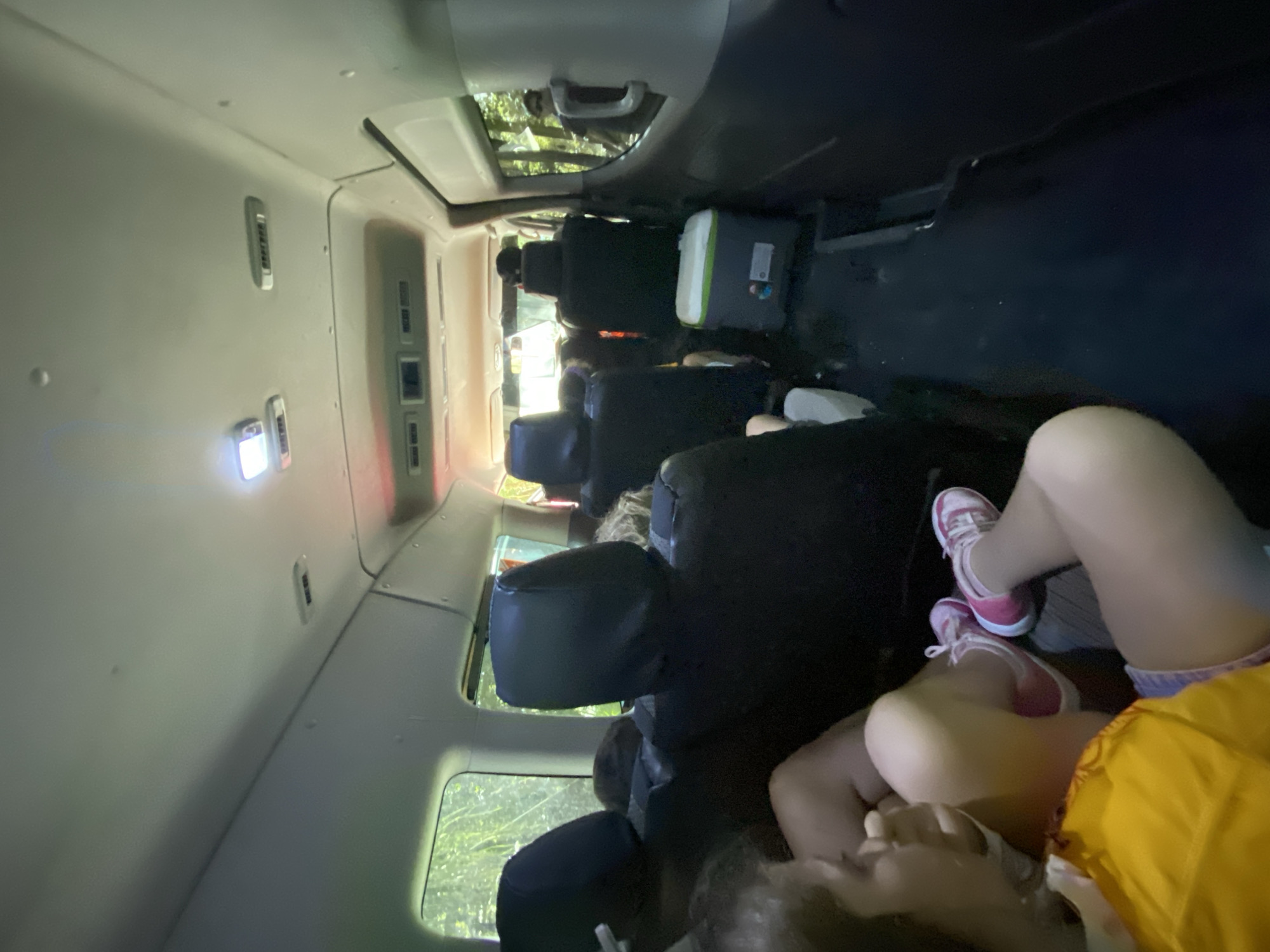 The interior of a passenger van is depicted, oriented with the front of the vehicle at the top of the image. A row of unoccupied black seats stretches from the midground toward the back, where a cooler with white and green accents is visible. Through the van's windows, foliage suggests a wooded area. In the foreground, a child's bare legs and pink and white sneakers are visible, along with a yellow garment. The child appears to be lying down. The photo was taken in a passenger van in Leona Vicario, Mexico.
