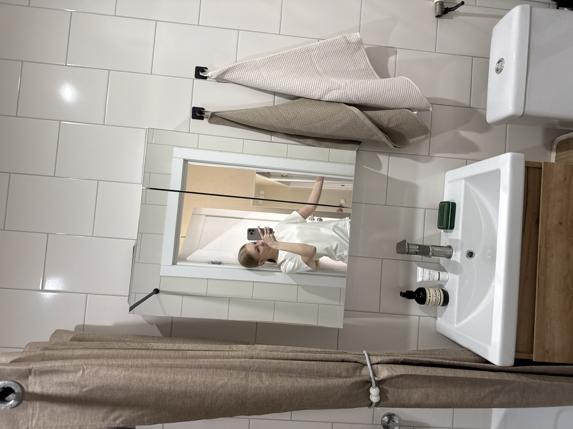 In a brightly lit bathroom, a young woman with blonde hair is taking a mirror selfie. She's wearing a white t-shirt and holding a smartphone in her right hand. The bathroom features white subway tiles on the walls. Two folded towels hang from black hooks mounted on the wall above the sink. To the right of the sink, a wooden cabinet is visible. The sink itself is white porcelain with a silver faucet. On the sink's edge, there are a few toiletries, including a small white tube and a dark glass bottle. A shower curtain, a neutral beige color, is drawn across part of the frame in the foreground. The overall setting suggests a modern and clean bathroom. The lighting indicates it is daytime. There are no obvious weather cues. The location is identified as Perm, Russia. There is some text visible on the dark glass bottle, but it is too small to be read clearly.