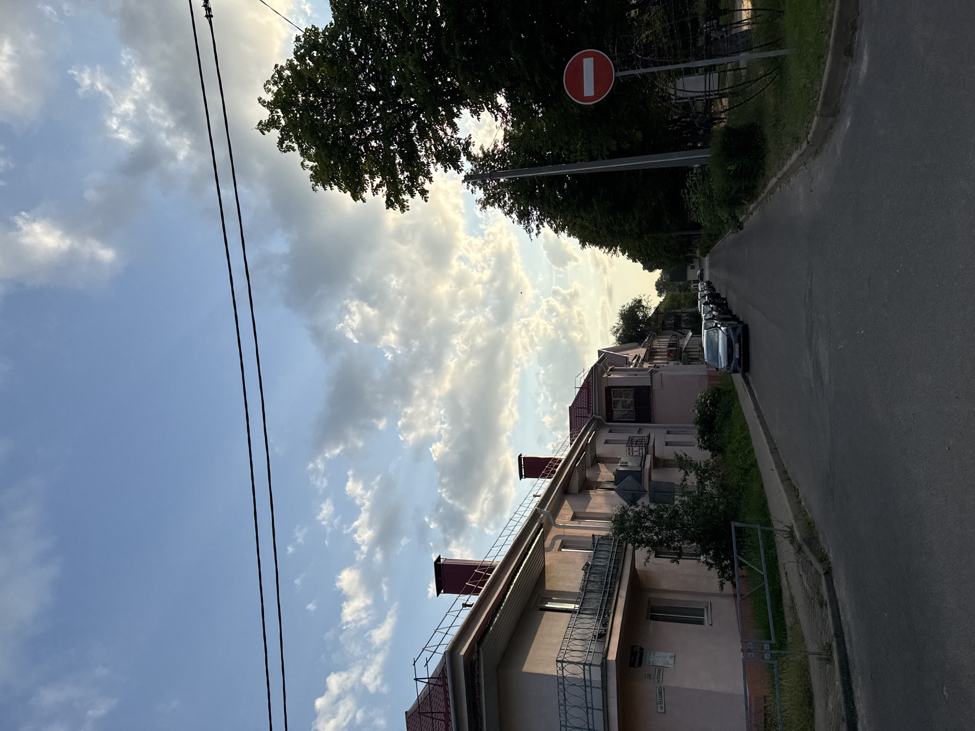 This image depicts a street scene in Zhodzina, Belarus, likely taken in the late afternoon or early evening, judging by the warm, diffused sunlight illuminating the clouds and the shadows. The sky is partly cloudy, with dramatic, illuminated cumulus clouds against a backdrop of blue.
The perspective is from a low angle, looking upwards and along a residential street. On the left side of the frame, the tops of trees and power lines are visible against the sky. To the right, a multi-story building with a pale pink facade dominates the mid-ground. The building has several balconies with decorative metal railings and visible rooflines with chimneys. Further down the street, more buildings and dense green trees line the road.
The street itself is paved and appears to be a quiet residential area. Several cars are parked in a line along the side of the road, suggesting a residential setting. A red and white "no entry" sign is mounted on a pole amidst the trees on the right side of the street, indicating a traffic restriction. There is some grass and vegetation along the sidewalk and around the buildings.
The overall mood is peaceful and serene, capturing a quiet moment in a suburban or town environment. The lighting suggests a pleasant, mild day. There is some faint text visible on a plaque on the building, but it is illegible.