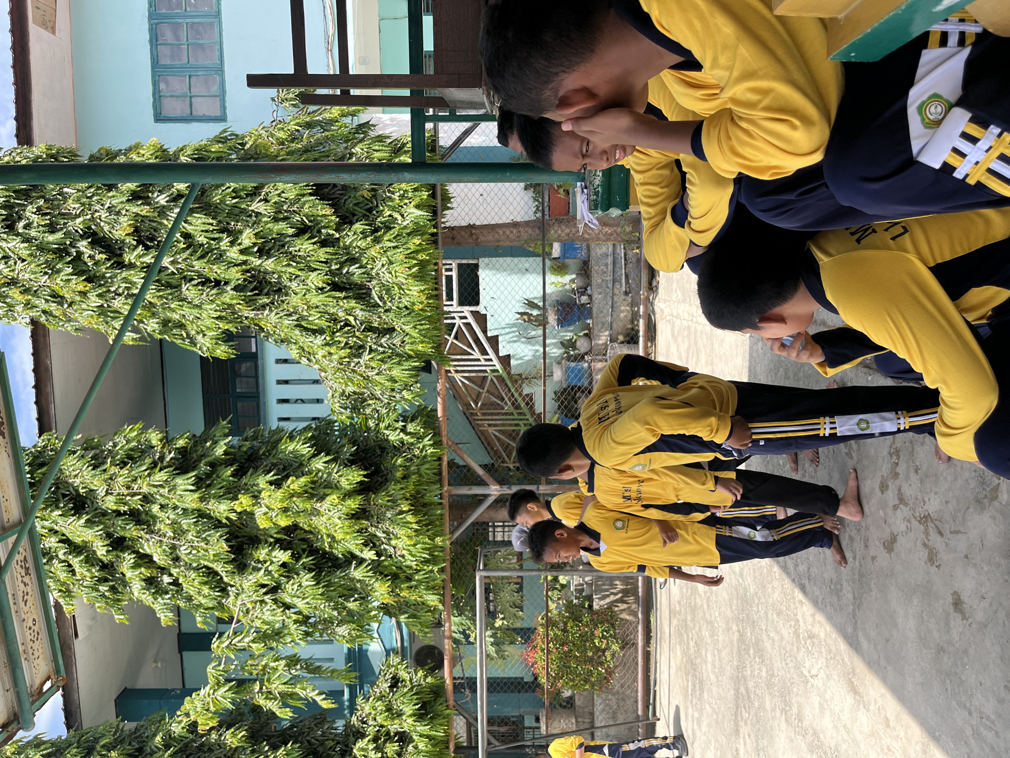 The image depicts a group of young boys, likely students, wearing matching yellow long-sleeved shirts with navy blue accents and navy blue pants with yellow and white stripes. They are standing and sitting outdoors in what appears to be a schoolyard. In the background, a light blue building with windows and an outdoor staircase can be seen, along with tall, leafy green trees. There is a fence surrounding the area. The ground is paved. Some of the boys are interacting with each other, with one boy touching another's face and another drinking from a bottle. The overall scene suggests a casual gathering or break time at a school in Lhokseumawe, Indonesia.