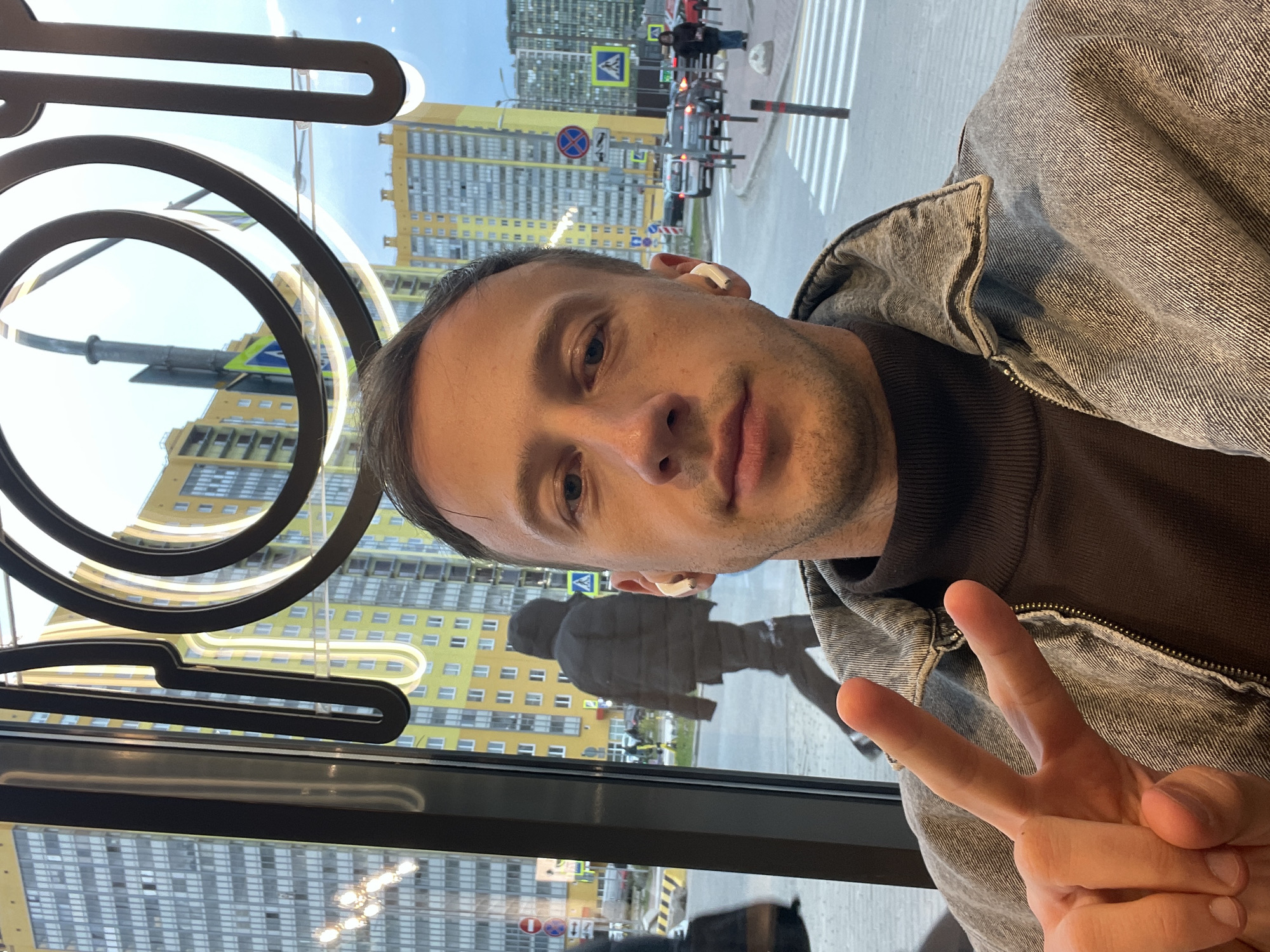 This media file is a rotated selfie taken by a man, likely indoors next to a large window overlooking an urban street scene. The man, with his head on the left side of the frame, has short dark hair and is wearing a brown high-necked top under a grey denim jacket. A white earbud is visible in his right ear. His left hand is raised towards the right, making a peace sign gesture. He looks directly at the camera with a neutral expression.
Through the window, which also reflects elements from inside the building, a bright daytime street scene is visible. Tall, multi-story residential buildings, one predominantly yellow and another grey, form the background. A street with a prominent zebra crossing is in the midground, with vehicles and a pedestrian visible. Traffic signs include a yellow triangle indicating a pedestrian crossing and a circular red sign denoting no stopping or parking. Reflections on the window reveal dark, abstract spiral shapes and a light fixture resembling a clothes hanger, along with the silhouette of another person. The sky outside appears bright blue and clear. The overall setting suggests a modern urban environment, consistent with a city like Saint Petersburg, Russia. There is no other discernible text.