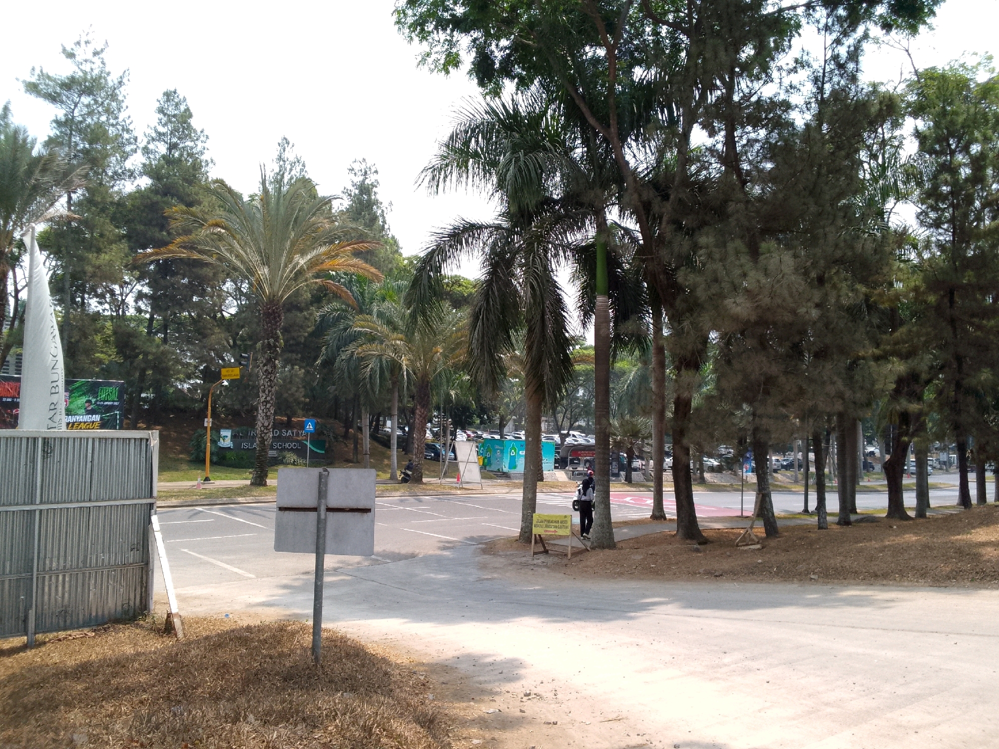 This image depicts an outdoor scene with a focus on a paved area that appears to be a parking lot or roadway, bordered by trees and vegetation. The foreground shows dry, brown grass and dirt, with a metal fence on the left side. A grey metal post with a blank white sign stands prominently in the mid-left. In the mid-ground, there is a paved area with white lane markings, and several palm trees and other trees line the background. A person wearing dark clothing and a backpack is standing near a wooden sign with text on it, on the right side of the paved area. In the far background, parked cars are visible, suggesting this is a public or commercial space. The presence of a sign that reads "RAHYANGAN LEAGUE" and another that mentions "ISLAMIC SCHOOL" indicates that the location is likely near an educational institution. Based on the contextual clues and the provided information, this scene is in Cipeundeuy, Indonesia. The overall lighting suggests a daytime setting, possibly with overcast skies or bright sunlight creating shadows.