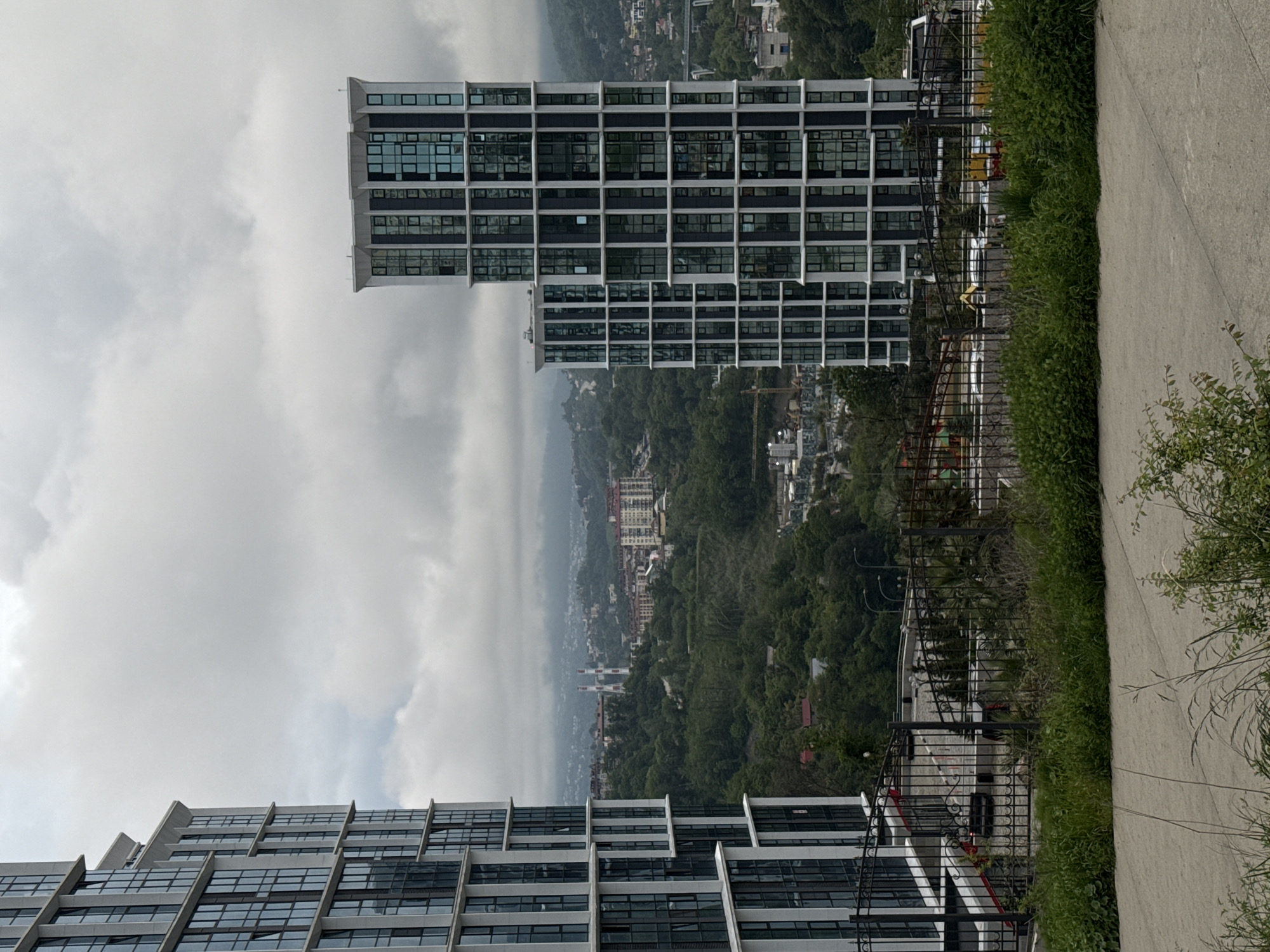 The image captures a scene from Sochi, Russia, featuring a view overlooking the city and the surrounding forested hills. Dominating the foreground are two modern buildings with a grid-like facade of windows and a reinforced concrete frame. The building on the left has a terraced structure. The building on the right is a high-rise with a perpendicular layout. Between the camera and the buildings, there's a concrete pathway bordered by a metal fence overgrown with vegetation, which gives the impression of a high vantage point. In the background, a low-lying cloud or mist obscures the mid-ground of the cityscape, partially concealing the city buildings, and mountains. The sky above is overcast and filled with clouds.