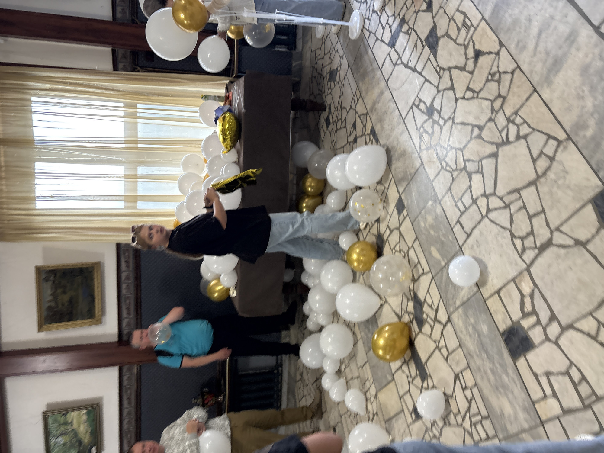 In this indoor scene, several people are gathered in a room decorated with numerous balloons. The balloons are predominantly white and gold, some are clear, and a few are patterned. They are scattered across the floor and arranged in clusters, suggesting a celebratory event like a birthday party or a holiday gathering.
The individuals visible are casually dressed. One person in the foreground is wearing a black t-shirt and jeans, looking towards the camera. Another individual in a blue t-shirt is also present. The floor is covered with a distinctive mosaic tile pattern in shades of beige and gray. In the background, a long table is visible, draped with a dark cloth. There are framed paintings on the wall, one of which depicts a landscape scene. The lighting suggests it is daytime, and the indoor setting provides no clues about the weather outside. The overall mood appears relaxed and festive. No visible text is present in the image. The location is identified as Zavod Mikhailovskii, Russia.