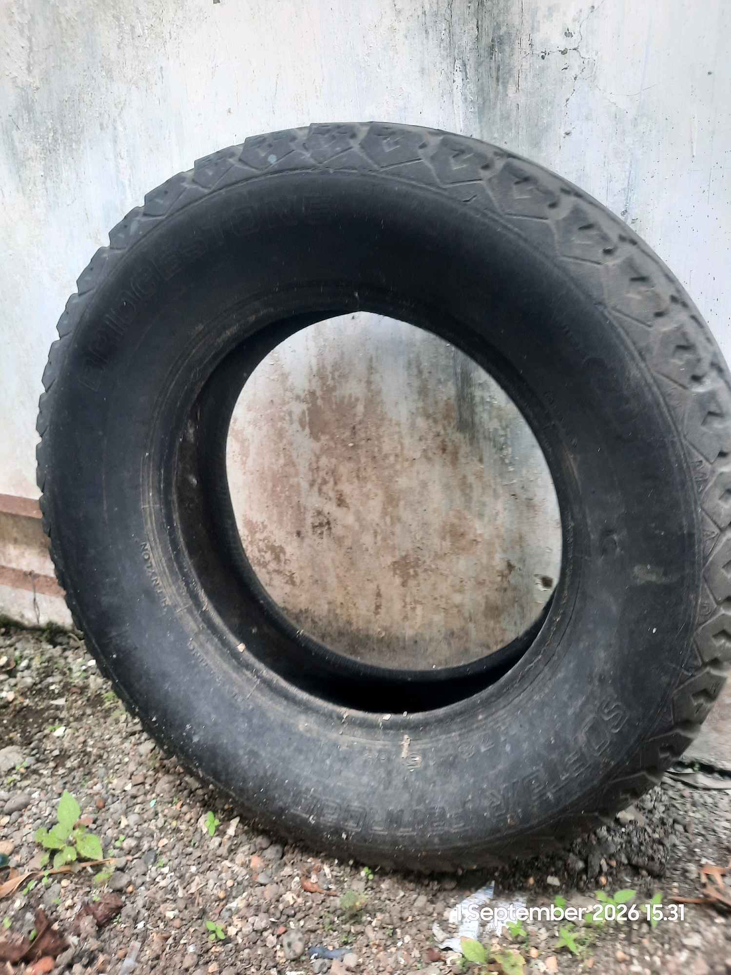 An old, worn tire lies on the ground against a textured, off-white wall. The tire has visible tread patterns and some text printed on its sidewall. The ground is covered in small stones and gravel, with a few green plants sprouting. A partial view of a reddish-brown structure is visible to the left of the tire. The scene appears to be outdoors. The image also contains a timestamp in the lower right corner: "1 September 2026 15.31". The location is Karanggeger, Indonesia.