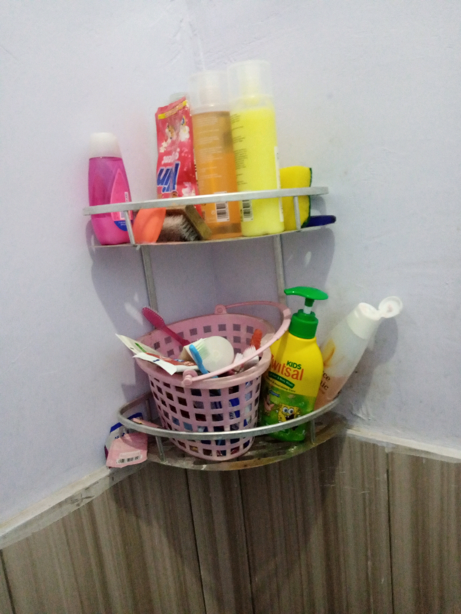 An indoor, eye-level, medium shot captures a corner shelf in what appears to be a bathroom in Sumbergirang, Indonesia. The shelf is made of metal and holds various toiletries. On the top shelf, from left to right, are a pink bottle with a white cap, a red package with cartoon characters, an orange bottle, a yellow bottle with a white cap, and a yellow sponge. A toothbrush and a brush are also visible. On the bottom shelf, a pink woven basket contains toothbrushes and other small items. To the right of the basket is a yellow pump bottle labeled "KIDS," with a green dispenser and an image of SpongeBob SquarePants. Next to it is a white bottle with a partially open white cap. The wall behind the shelf is white and slightly textured, while the lower portion of the wall features light brown tiled paneling. The lighting is dim, creating shadows on the wall.