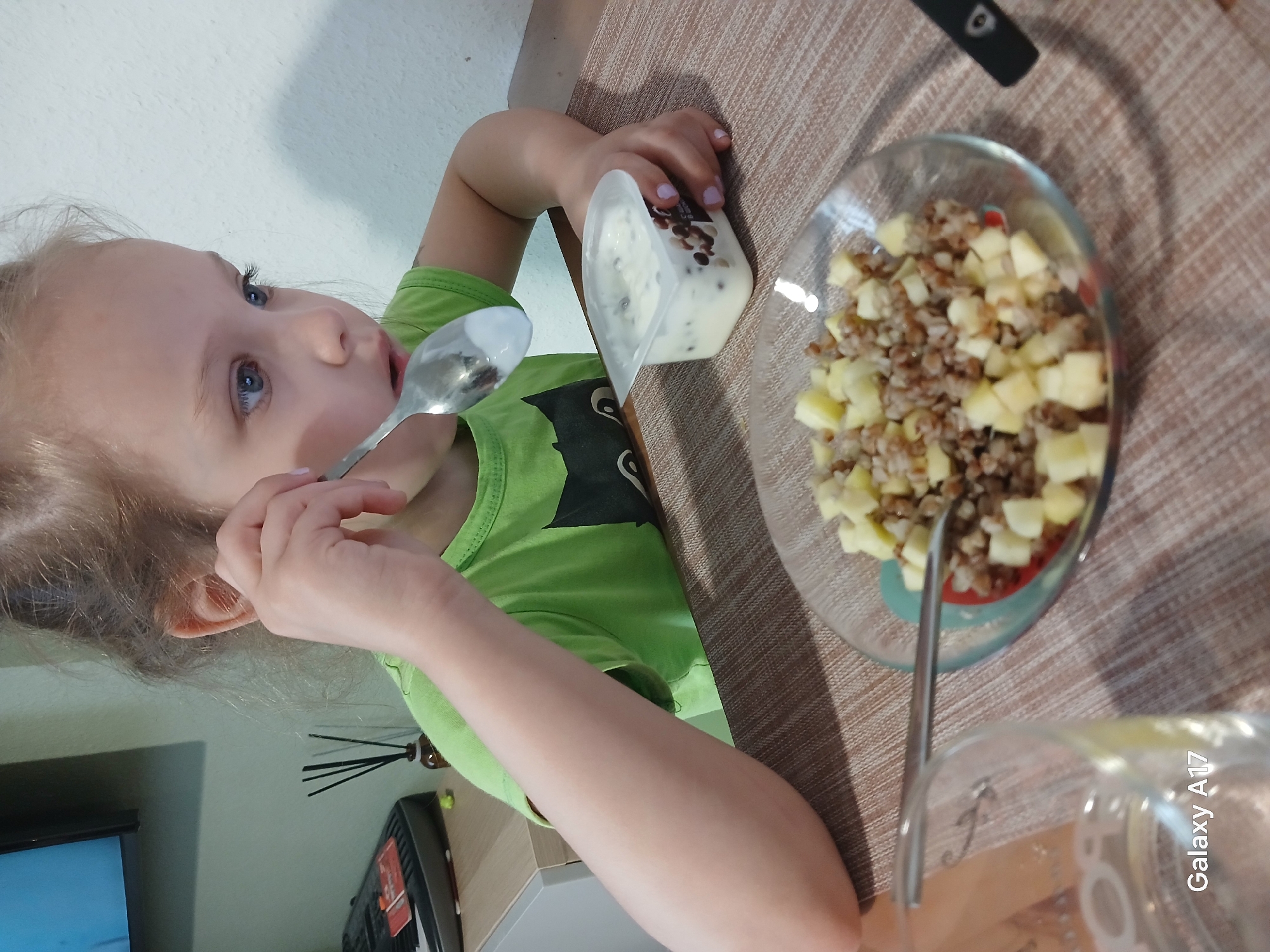 A young child with blonde hair and blue eyes, wearing a green shirt with a black design, is eating at a table. The child holds a spoon to their mouth. Next to the child is a small, clear container of what appears to be white yogurt with dark speckles. A clear glass bowl with chopped apples and a brown grain, possibly buckwheat, is also on the table. A spoon rests in the bowl. The table is covered with a brown patterned tablecloth. In the background, partially visible, are a TV, a keyboard, and an air freshener. "Galaxy A17" is written in the lower right corner of the image. The specific location in Yekaterinburg, Russia, cannot be determined from the given image.