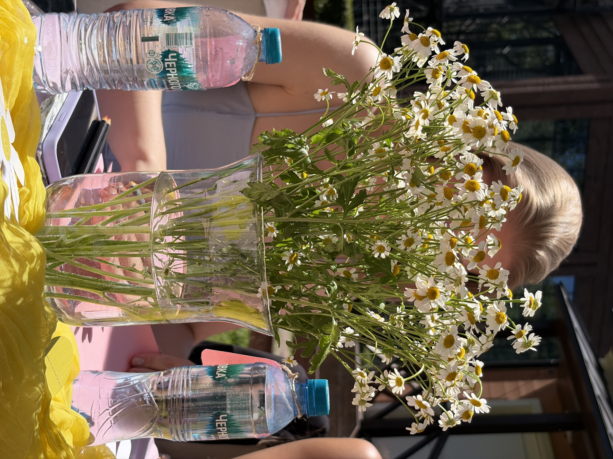 A bouquet of daisies is centered in a clear glass vase filled with water, placed on a pink surface. Two bottles of water, one with a pinkish liquid and the other clear, are positioned to the left and right of the vase, respectively. The bottle on the left has a blue cap and a label with Cyrillic script. The bottle on the right also has a blue cap and a label with Cyrillic script and a pink tag. A person with blonde hair is partially visible behind the bouquet, their face obscured. In the foreground, a yellow fabric is draped across the pink surface, with a white object that appears to be a napkin with yellow cutlery placed on it. The background shows a building with a wooden facade and dark window frames, suggesting an outdoor setting. The overall impression is a picnic or an outdoor gathering. The presence of Cyrillic script on the water bottles indicates a location in Russia, and the mention of the city of Vidnoye in the prompt confirms the location.