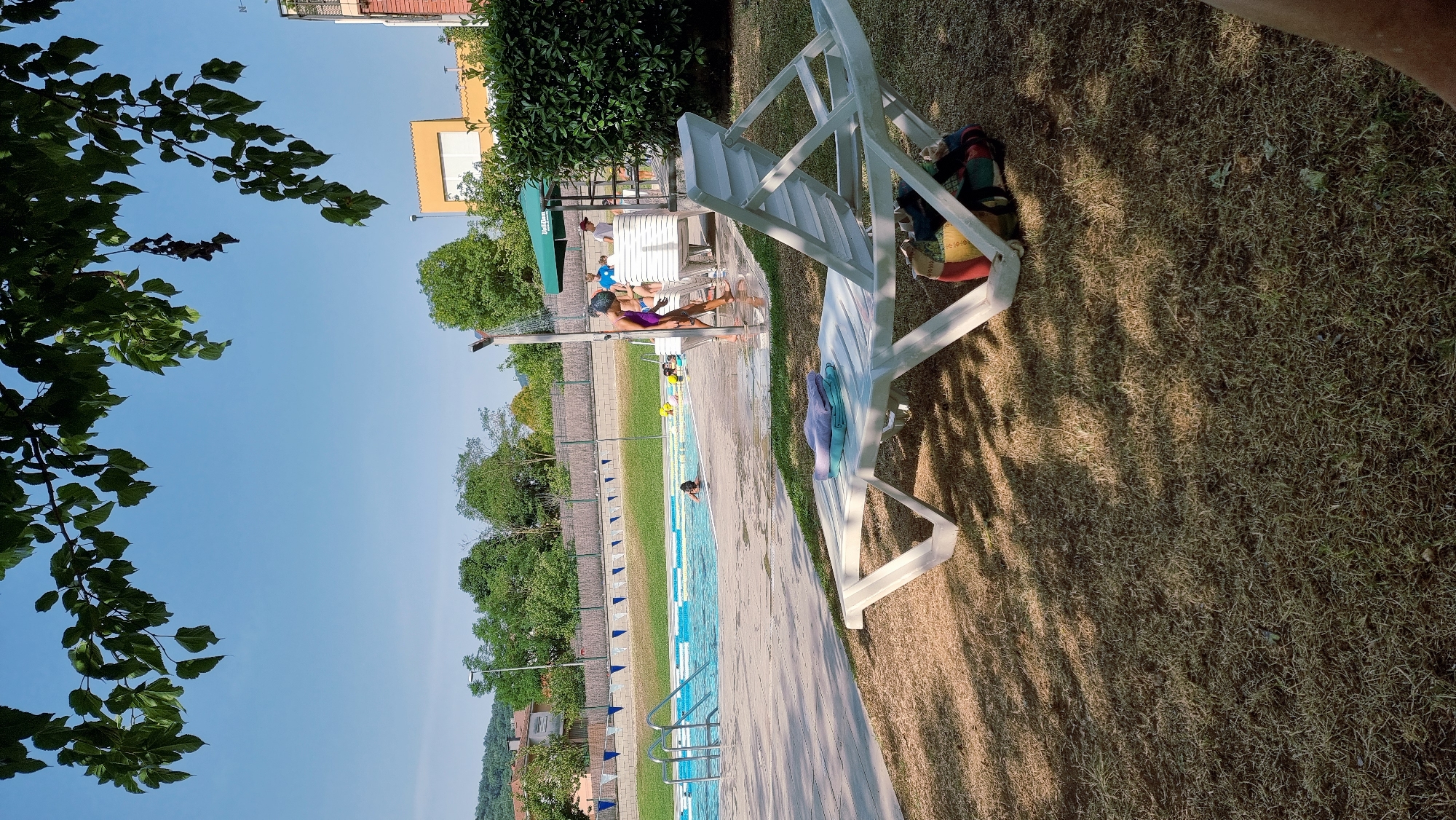 The image captures a scene at an outdoor swimming pool in Sant Joan Les Fonts, Spain, on a clear, sunny day. In the foreground, a white plastic lounge chair is placed on a dry, grassy area, casting a shadow. The chair has folded towels draped over it. Beyond the chair, the pool area is visible, with blue and white lane markers in the water. Several people are present: some are in the pool, one person, appearing to be a young girl in a purple swimsuit, is standing under an outdoor shower, and others are milling about near a white structure. A green awning is visible above the white structure. In the background, trees and buildings are scattered across a hilly landscape under a bright blue sky. The overall impression is one of a recreational area enjoying pleasant weather.