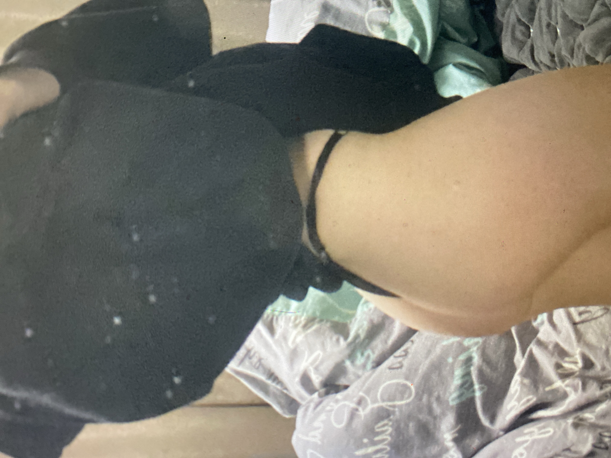 The image displays a partial view of a person in an indoor setting, likely a bedroom, situated in Ternopil, Ukraine. The central subject is a light-skinned person, whose arm or shoulder area is visible on the right side of the frame. A thin black strap, possibly from a bra or top, wraps around the neck or upper shoulder area. The person is wearing black clothing, covering the left and upper parts of the body, which appears to be a textured fabric with some white specks, possibly lint or dust.
Below and around the person, there is a pile of fabrics, resembling unmade bedding or discarded clothes. One piece of fabric is a light blue or mint green color, while another is light grey or lavender. The grey/lavender fabric features decorative white text in a cursive style; parts of words like "dreams" and other letters such as "h," "a," "li," and "s" are partially discernible, suggesting a pattern like "Sweet Dreams." The overall lighting is soft, but the specific time of day is not evident. The image itself appears to be a photograph of a screen, exhibiting a visible pixel grid and some graininess. No specific activity is depicted other than the static pose of the person.