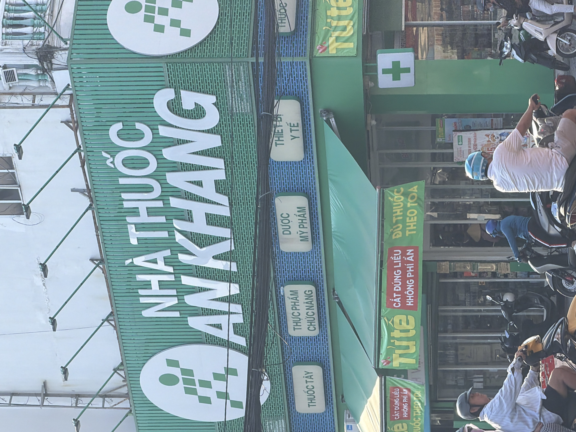 The image captures a street view in Nha Trang Ward, Vietnam, focusing on a pharmacy called "NHÀ THUỐC AN KHANG". The pharmacy's facade features a prominent green and white sign with its name in large white lettering. Below the main sign, smaller signs indicate different product categories: "THUỐC TÂY", "THỰC PHẨM CHỨC NĂNG", "DƯỢC MỸ PHẨM", "THIẾT BỊ Y TẾ", and "THUỐC". The pharmacy also has green awnings covering the storefront. A green sign with a white cross, a common symbol for pharmacies, is visible on the right side of the building.
In the foreground, several people on motorcycles are depicted. On the left, a person wearing a light-colored striped shirt and dark shorts rides a yellow and black motorcycle. In the center, a person in a light pink shirt and light-colored shorts, wearing a blue helmet, rides a black motorcycle. On the right, a person in a blue shirt and dark pants, also wearing a helmet, rides a dark-colored motorcycle. Additional motorcycles are parked or in motion around them. The environment suggests a bustling urban street with commercial establishments.
