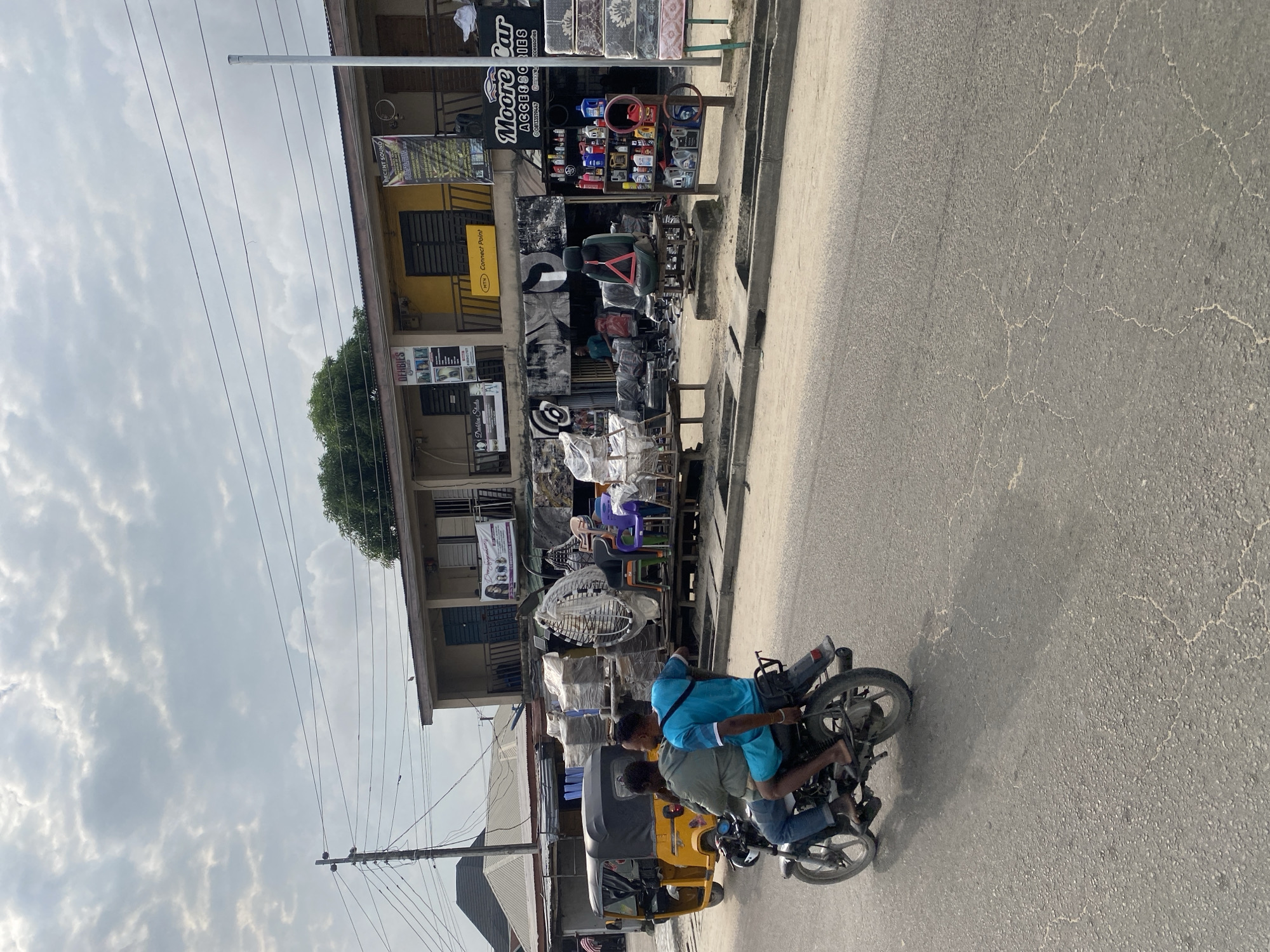 The image depicts a street scene in a bustling urban environment, possibly in West Africa. In the foreground, two men are riding a motorcycle. The man in the back is wearing a blue t-shirt and shorts, while the man in front is wearing a green shirt and jeans. The motorcycle is moving on a paved road with visible cracks.
Behind the motorcycle, a yellow tricycle, commonly known as an "achaba" or "keke," is parked. The buildings lining the street are commercial establishments. One building is painted yellow and has multiple shops with signs. A prominent sign reads "Moore Car ACCESSORIES." Another sign says "DEHBIES." Various items, including car accessories, chairs, and stacked mattresses, are displayed outside the shops. The sky is overcast with clouds. The overall impression is a lively street with active commerce.