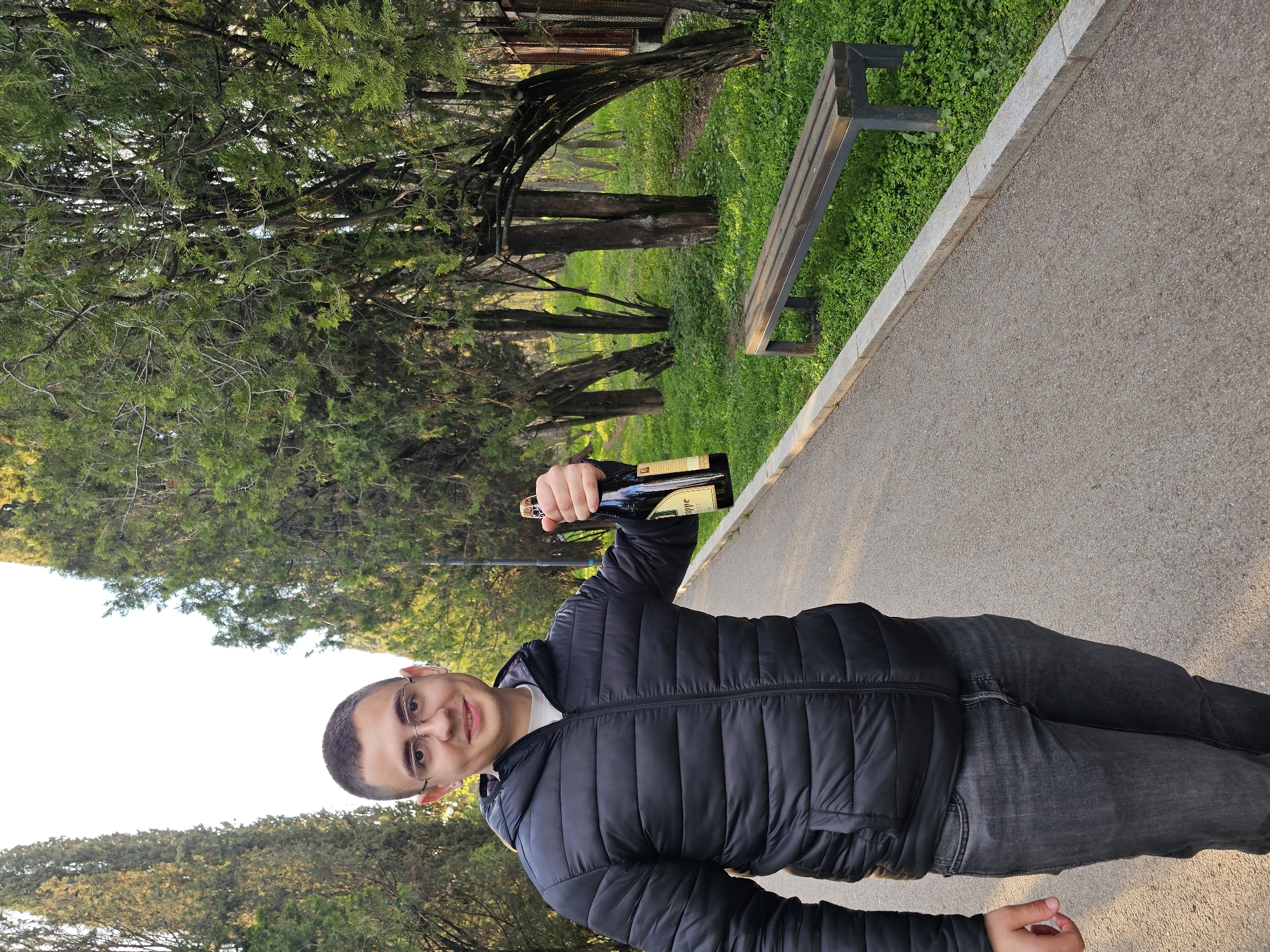 A man stands on a paved path in what appears to be a park or green space, holding a bottle of champagne in his right hand. He is wearing a black puffer jacket and grey jeans. The area around him consists of trees and some grass patches. A bench is visible next to the path. The location appears to be a park-like setting, with the path suggesting a recreational area for walking or jogging. Based on the characteristics of the flora and general layout, it is possible that the location is within the city of Stara Zagora, Bulgaria.