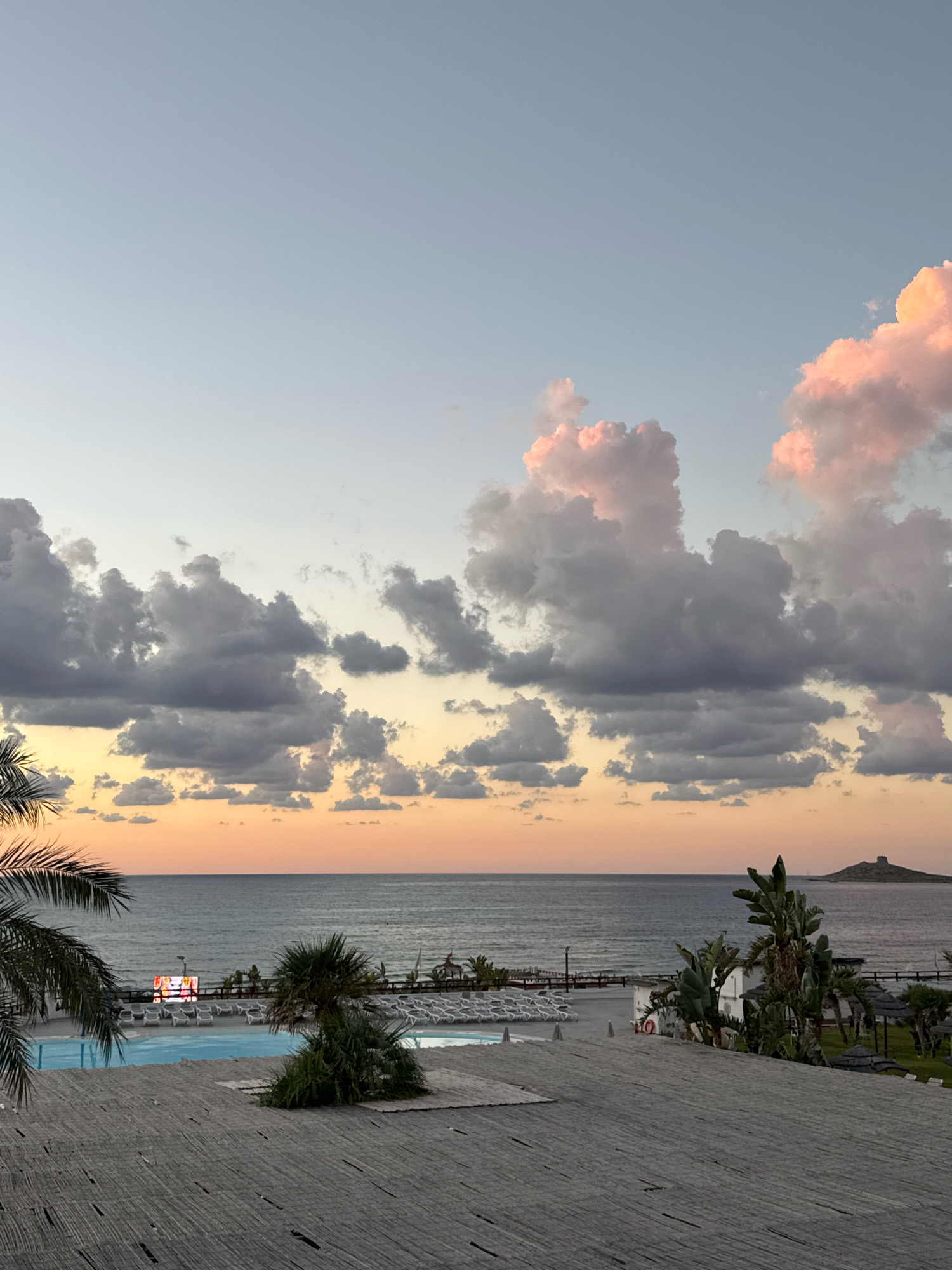 The image depicts a coastal view with a resort in the foreground. The sky transitions from a pale blue at the top to a warm orange and yellow gradient near the horizon, punctuated by scattered clouds. Some clouds are tinged with pink, suggesting a sunset or sunrise.
In the mid-ground, a calm sea stretches towards the horizon. Along the coastline, there are rows of white lounge chairs arranged around a swimming pool. Palm trees and other tropical vegetation are visible, along with some resort buildings. A large outdoor screen displays colorful images. In the far distance, a small, conical island with a structure on top is visible.
The foreground is dominated by a weathered, grey wooden deck, constructed from parallel planks. Palm fronds are visible in the lower left corner, adding to the tropical atmosphere. The overall scene suggests a peaceful, resort setting in a coastal location. The presence of lounge chairs, a pool, and palm trees indicates a place designed for relaxation and leisure. The distant island hints at the specific geography of the location, which is identified as Capaci, Italy.