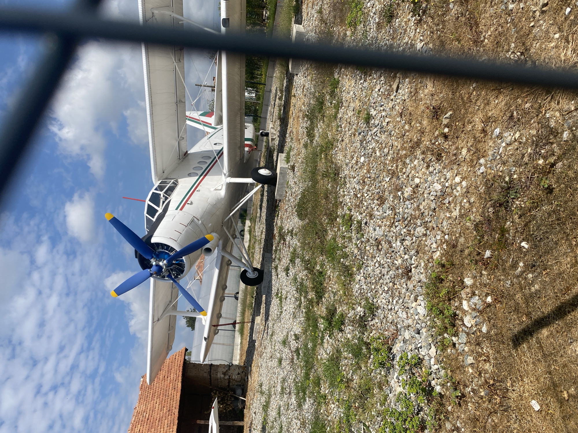 Observed from behind a barrier (possibly a fence), the image depicts a white biplane with blue and yellow propeller blades. The aircraft displays green and red stripes on its fuselage and wings. It is situated on a gravel surface interspersed with dry grass and some green vegetation. In the lower left corner, a portion of a brick structure is visible. The sky overhead is blue with scattered white clouds. The image is taken in Kapatovo, Bulgaria.