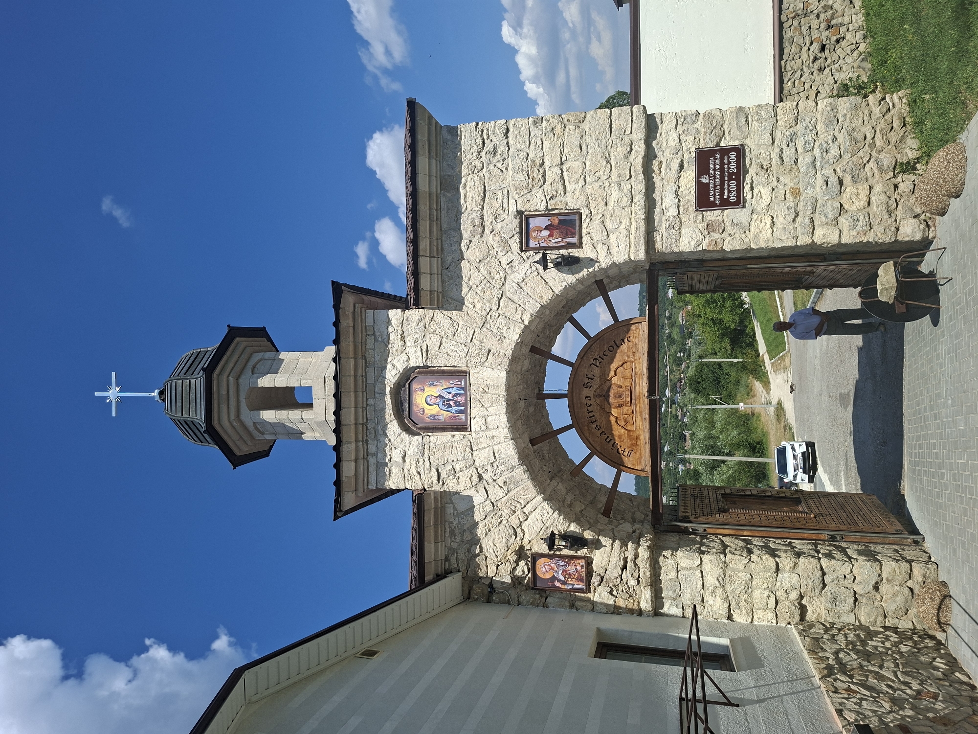 The image shows the entrance gate to a monastery in Condrița, Moldova. The gate is made of stone and has a wooden archway with religious icons above and an inscription in Romanian. A cross adorns the top of the gate tower, which is capped with a dark dome. To the left of the gate is a white building with a stone base. The entrance to the monastery is open, revealing a paved courtyard and a road leading away from the gate. A white car is parked on the road, and a man in a light blue shirt and grey pants stands near the gate. In the background, there are trees and a village with houses. The sky is blue with white clouds. A sign on the right side of the gate reads "MANASTIREA CONDRITA - SFANTU IERARH NICOLAE" and lists the opening hours as "08:00 - 20:00".