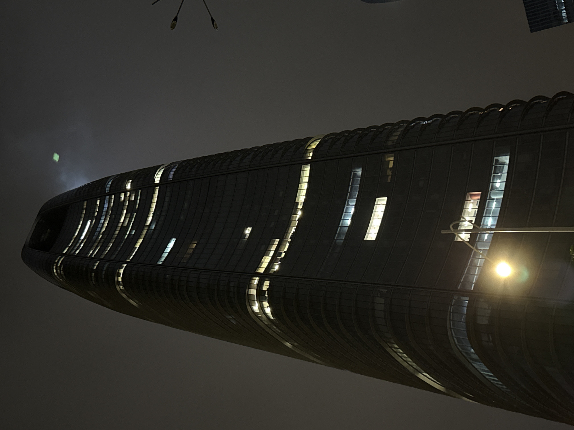 The image shows a close-up, low-angle shot of a modern, dark skyscraper in Haizhu District, China, at night. The building has a curved, segmented facade with numerous windows, many of which are illuminated with warm light, suggesting occupancy. The sky is dark and overcast, with a faint, square green light visible in the upper right quadrant, possibly a drone or a distant light source. A streetlamp in the foreground casts a bright yellow glow, its pole extending upwards and partially obscuring the lower section of the building. The overall impression is of a sleek, contemporary urban environment at night.