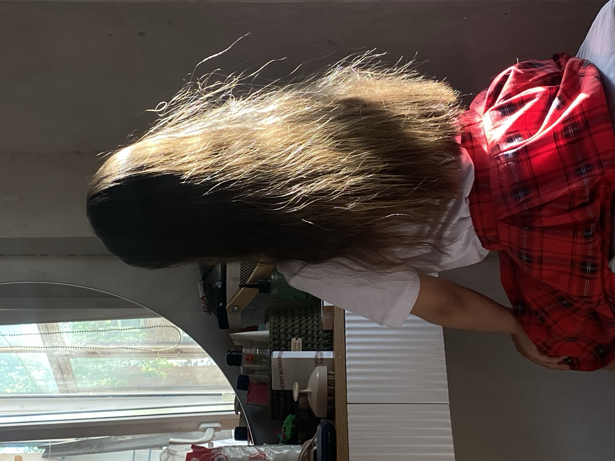 A person with long, light brown hair is sitting indoors. They are wearing a white t-shirt and a red plaid skirt. In the background, there is a window with a view of greenery and a shelf with various items, including bottles and a brush. Given the reference to Votkinsk, Russia, the scene is likely located in that city.