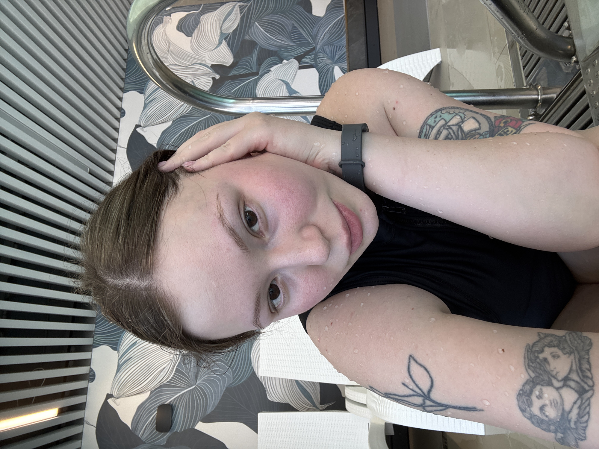 A woman is photographed from a close, high-angle perspective. She is likely at a pool or spa, indicated by the water droplets on her skin and the wet appearance of her dark, sleeveless top. She has fair skin, with a rosy complexion on her face, brown hair, and light-colored eyes. A gray fitness tracker or smartwatch is visible on her wrist. Her left arm has two tattoos: one appears to be a flower branch and another depicts two human figures in dark ink. A patterned surface of blue and white stylized leaves and a metal railing, likely part of pool access, are in the background. The location is potentially Chaikovsky, Russia, based on the prompt context.