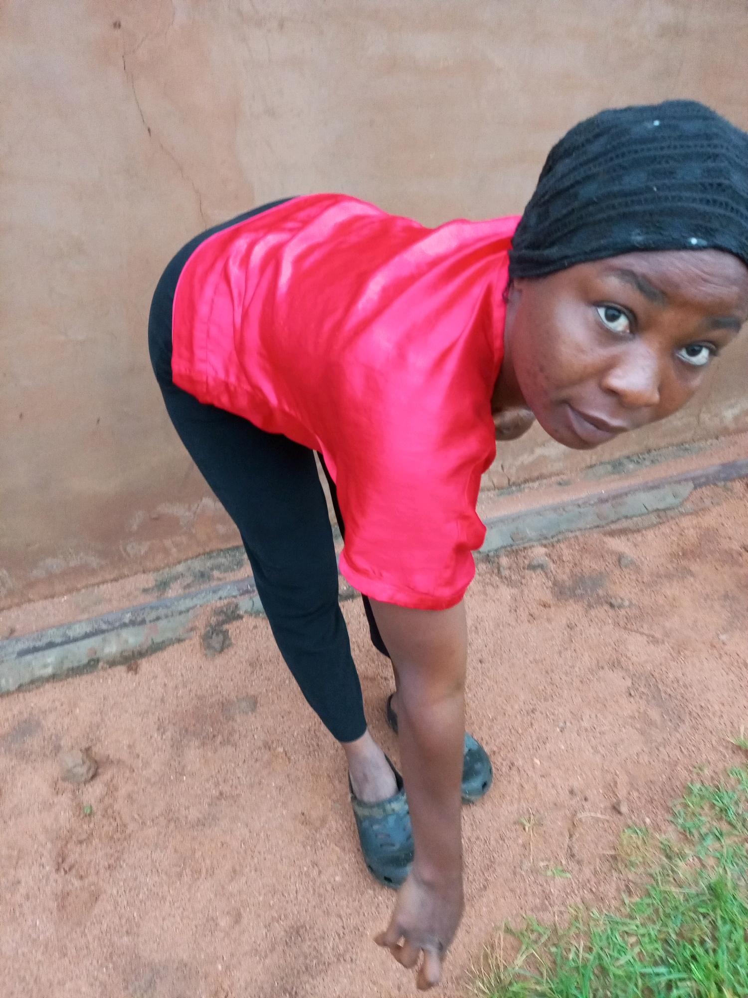 The image depicts a person wearing a black headscarf, a vibrant pink shirt, and black trousers. They are also wearing black clogs. The person is bent over, with their gaze directed towards the camera. The background consists of a textured, light brown wall and a reddish-brown dirt ground. Sparse green grass is visible in the lower right corner of the frame. The scene appears to be outdoors, with the wall suggesting a building structure. The overall impression is of an informal, everyday setting.