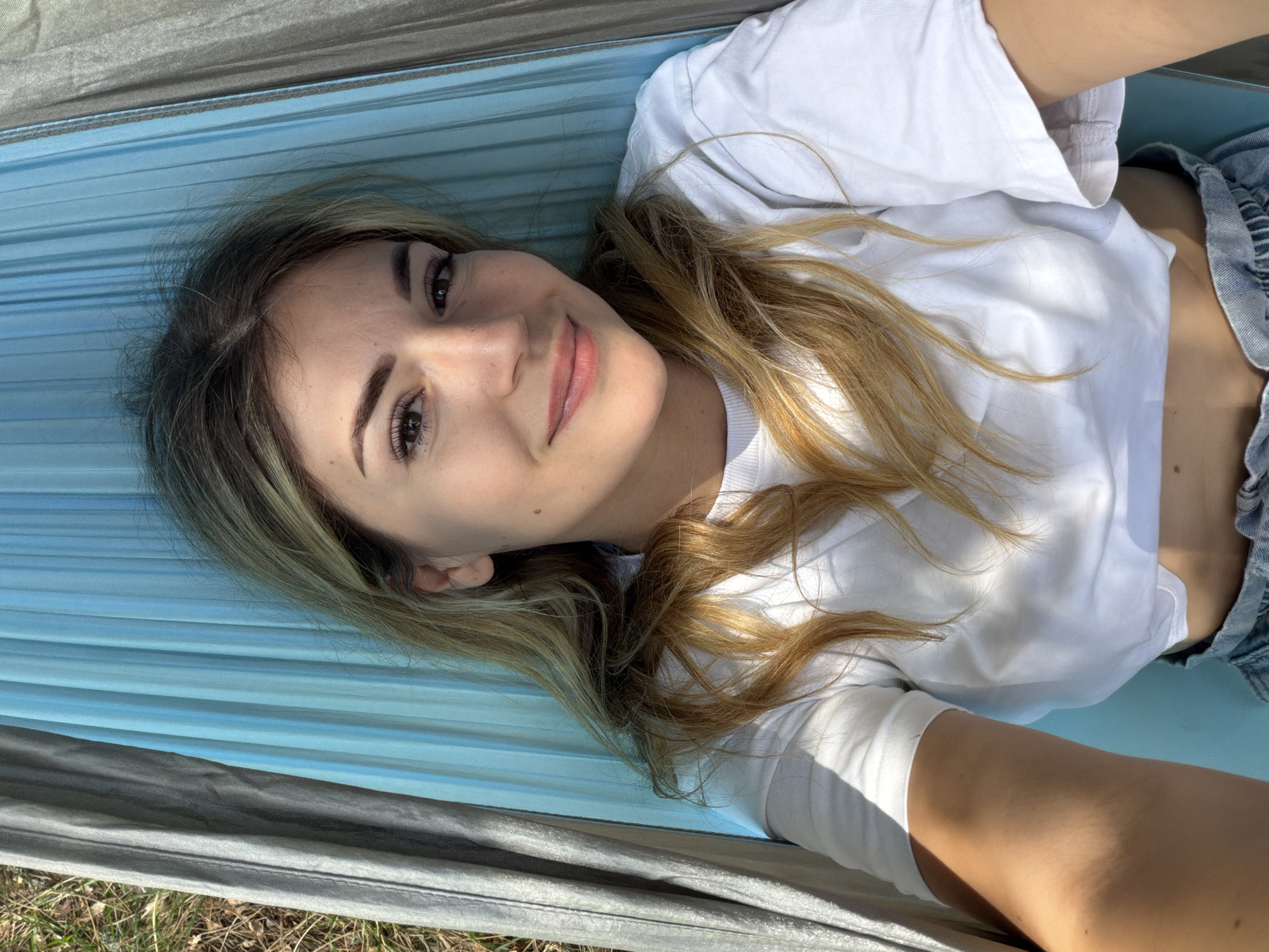 A young, light-skinned woman with long, blonde and light brown hair lies in a hammock in an outdoor setting in Mali Bostur, Montenegro. She is wearing a white short-sleeved crop top and light blue denim shorts. Her head is positioned towards the top of the frame, and she looks directly at the camera with a subtle smile. Her eyes are brown, and her eyebrows are dark and well-defined. A small mole is visible on her right cheek. Strong natural light illuminates her face and right arm, casting some shadows. The hammock is composed of light blue and dark grey sections, made of a synthetic fabric textured with vertical folds. Dry grass and foliage are visible behind the left edge of the hammock, confirming an outdoor environment.
