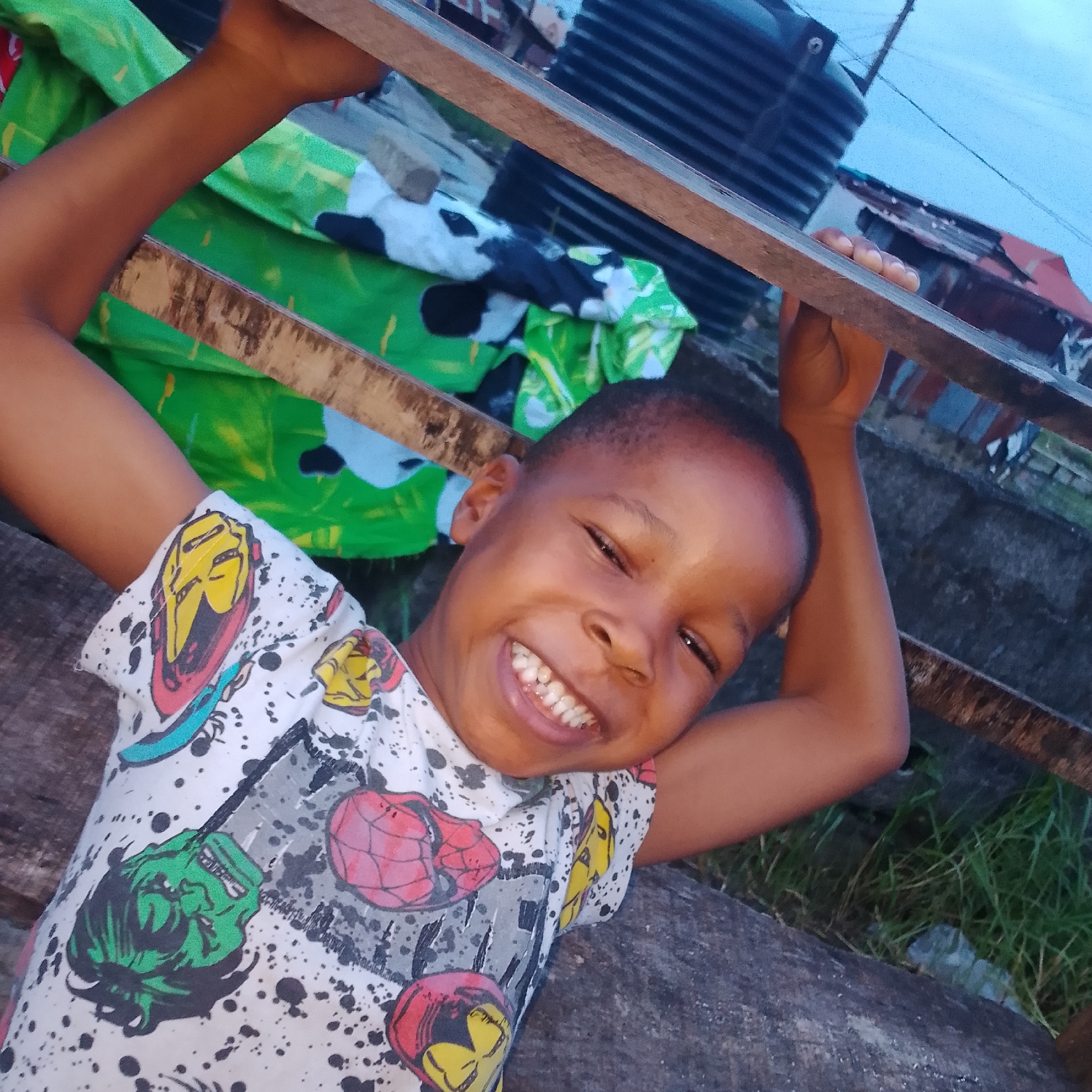 In Ado, Nigeria, a young boy with dark skin and short black hair is pictured outdoors, leaning against a wooden railing. He is wearing a white t-shirt with colorful comic book characters printed on it, including what appear to be superheroes. The boy is smiling broadly, with his teeth showing. His arms are raised and holding onto the railing above him. In the background, there is green fabric draped over something, and hints of corrugated metal roofing and buildings, suggesting a semi-urban or rural setting. The overall lighting indicates it is likely late afternoon or early evening, with a pale blue sky visible.