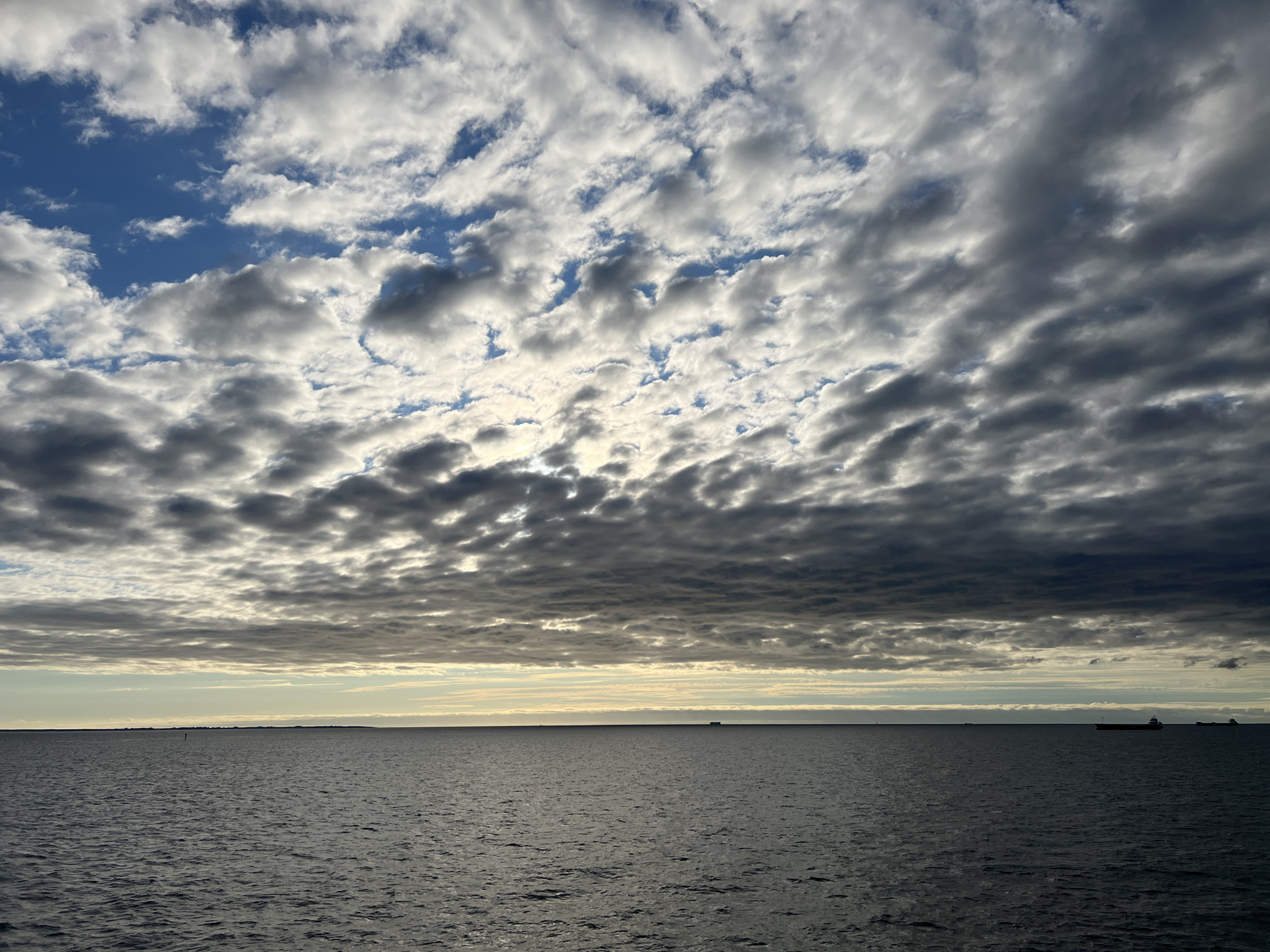 The image depicts a wide expanse of the Baltic Sea under a dramatic sky. The sea is a deep, dark gray with gentle ripples, suggesting a calm but overcast day. The horizon is a sharp, thin line separating the water from a band of pale, yellowish-green sky, indicative of either sunrise or sunset, or a break in the clouds. Above this band, the sky is filled with a complex tapestry of clouds. Some are bright white and fluffy, catching the light, while others are darker, more dense, and cast shadows, creating a textured, layered effect.
In the distance, along the horizon, several dark silhouettes of ships are visible. One larger vessel, a cargo ship or tanker, is prominent on the right side of the frame. Other smaller dots suggest further vessels or buoys. The overall scene evokes a sense of vastness and the power of nature, with the human presence marked only by distant maritime activity.
The location is the Bay of Gdańsk, visible from the coast of Gdynia, Poland. The perspective is from sea level, likely from a boat or the shore, looking out towards the open water and the sky. The lighting suggests it is either early morning or late evening, with the sun positioned low on the horizon, partially obscured by clouds.