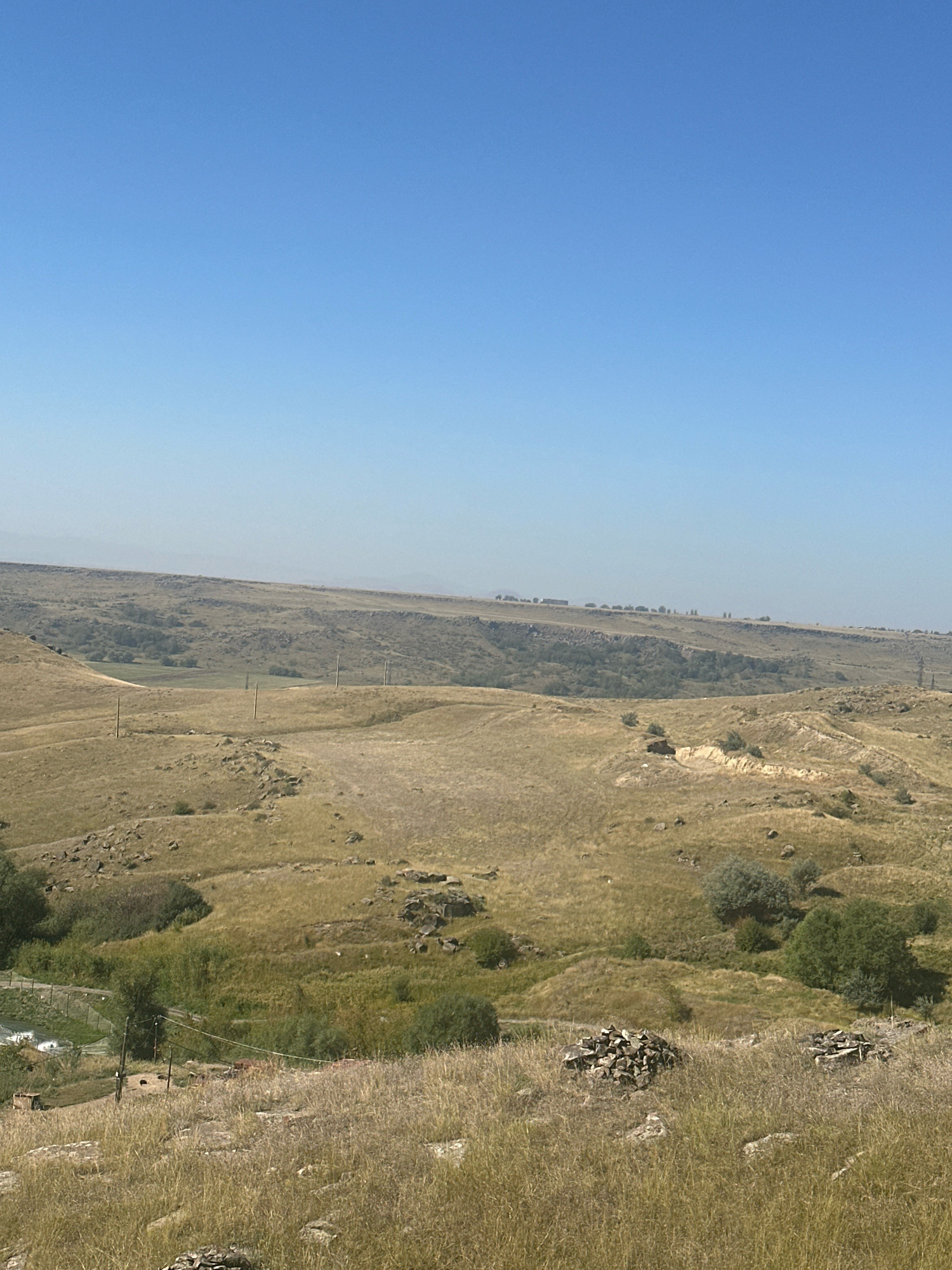 The image shows a wide, hilly, dry landscape under a clear blue sky. The terrain is predominantly covered in dry, yellowish-brown grass and sparse, low-lying green bushes and trees. Numerous large rocks are scattered across the landscape. In the middle ground, there are gently rolling hills and some evidence of human presence, including telephone poles, a faint road or path, and what appear to be small structures or ruins on some of the hillsides. In the far distance, the horizon is hazy, with faint outlines of what could be more hills or distant buildings. In the foreground, the dry grass is taller and more dense, with more rocks and two distinct piles of stones. A small fenced area with some green vegetation and a body of water is visible in the lower left quadrant. The overall impression is of a rural, arid environment. This landscape is consistent with the outskirts of Gyumri, Armenia, which is known for its hilly terrain and somewhat dry climate.