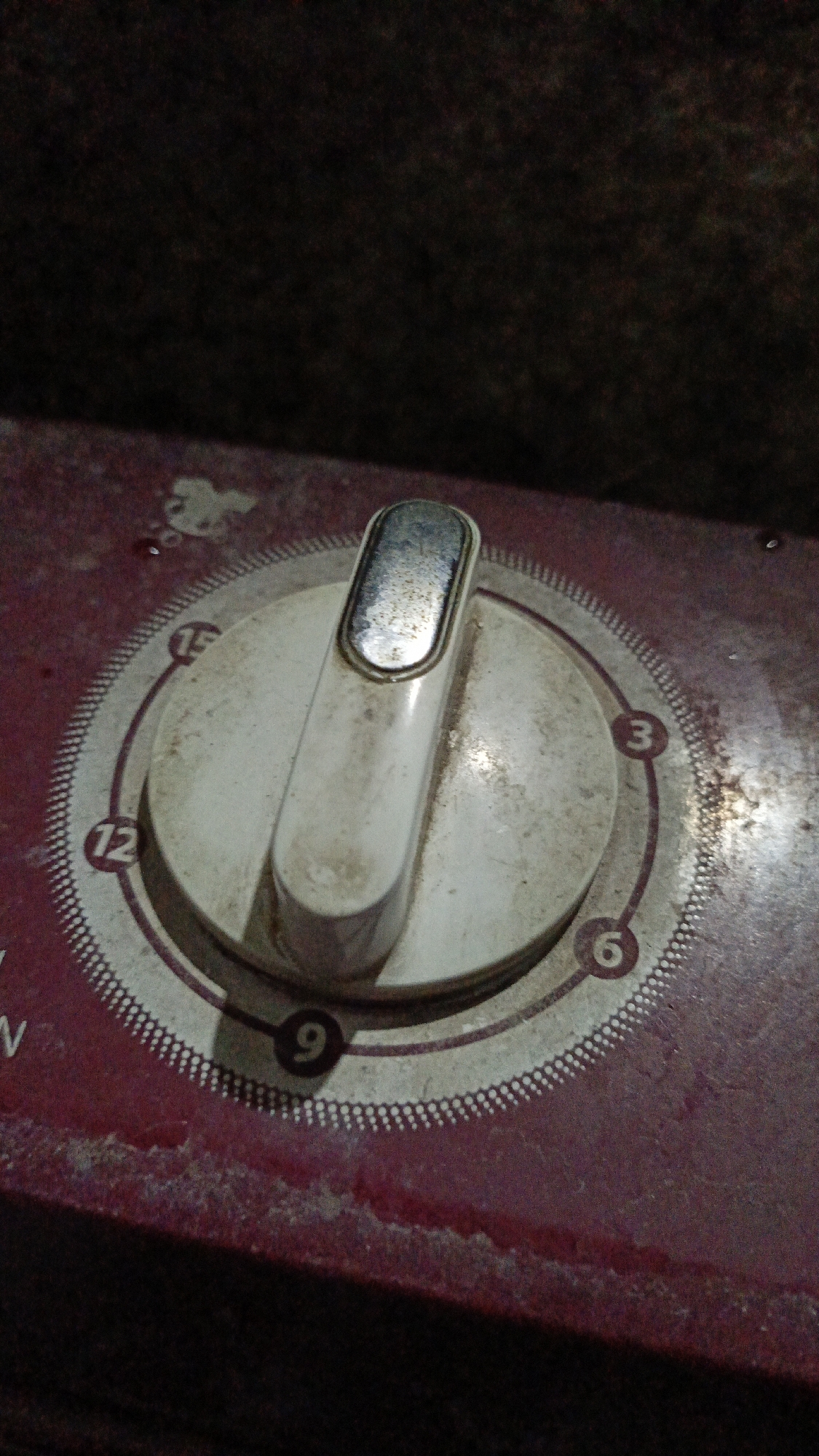 The image shows a close-up of a vintage rotary dial on a dusty, maroon-colored appliance. The dial has numbers 3, 6, 9, and 12 clearly visible, with an indicator knob positioned slightly past the 12. The dial itself is off-white and worn, with some scratches and dirt. The appliance's surface appears to be made of plastic or painted metal and shows signs of age and wear, with some chipped edges. The background is dark and out of focus, suggesting an indoor setting with low lighting. There are no people visible in the image. The scene depicts a detail of an aging electronic device, likely a timer or a control panel.