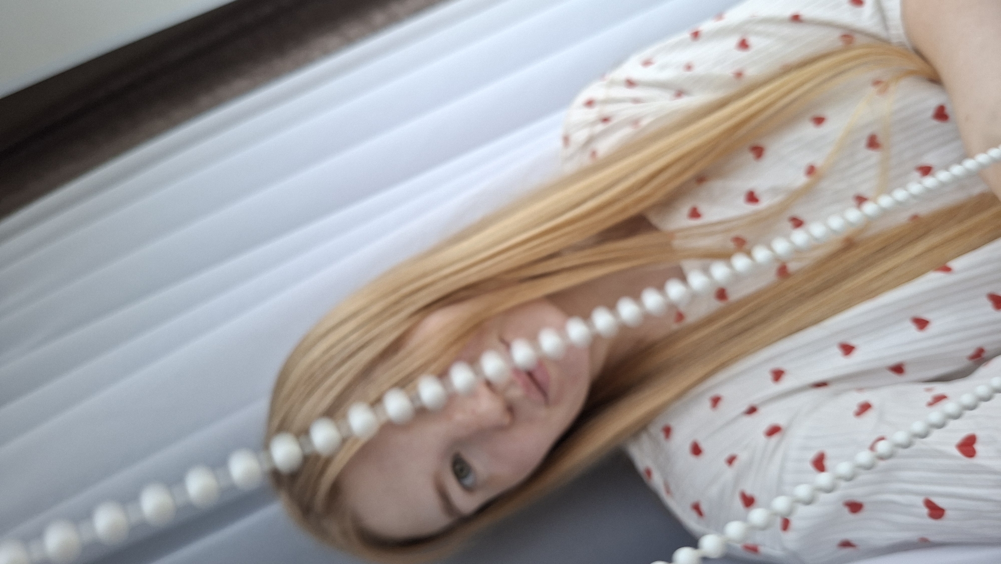 A young woman with long blonde hair, wearing a white top with red hearts, is pictured indoors, possibly in an apartment in Chaikovsky, Russia. She is partially obscured by a string of white beads hanging in front of her face. Behind her, white vertical blinds create a backdrop. The lighting suggests an indoor setting, possibly near a window.