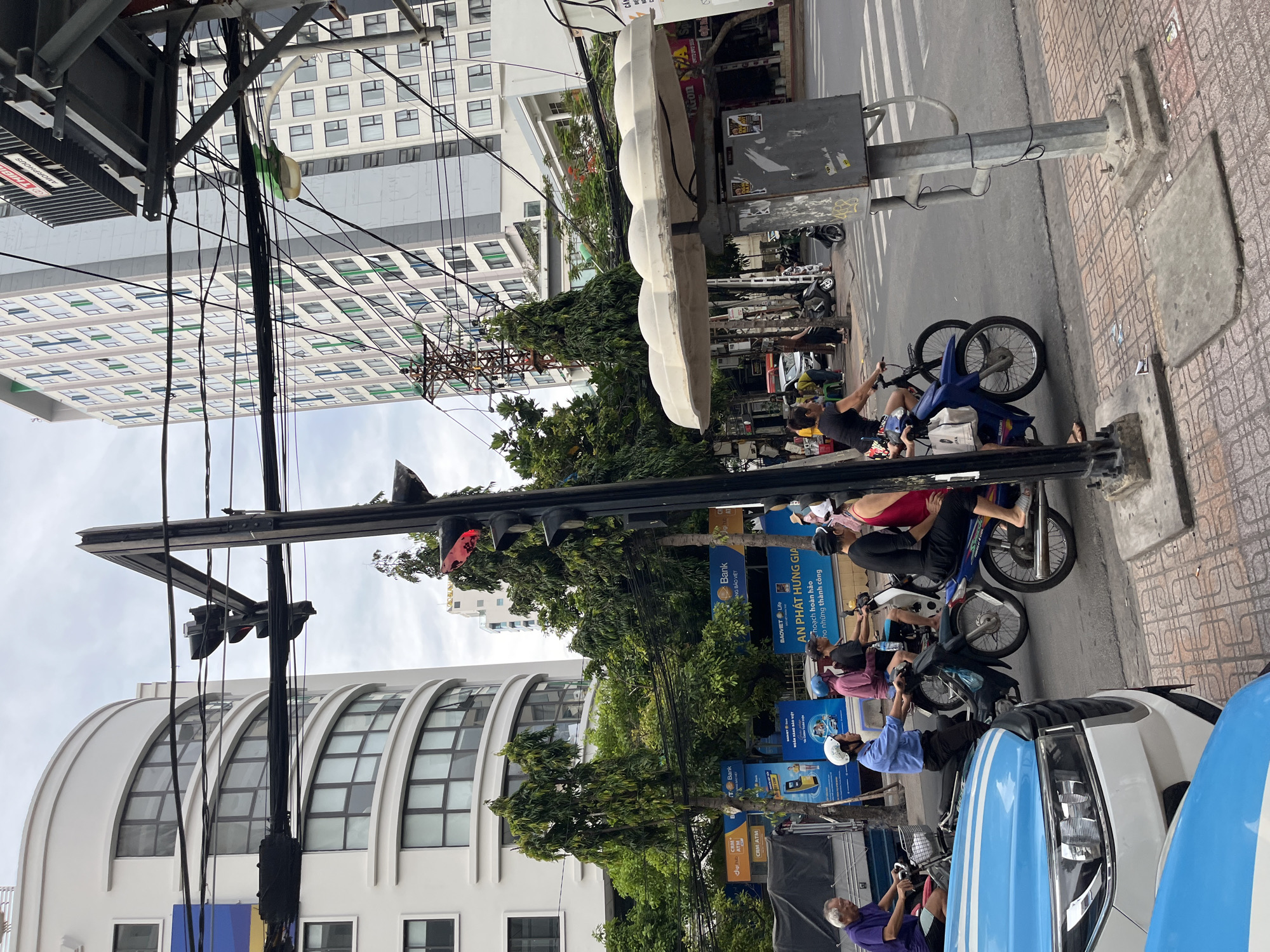 The image shows a street scene in Nha Trang, Vietnam, with a focus on a traffic light and several people on motorbikes. A traffic light signal is visible with a red light illuminated. Several individuals are riding motorbikes in various directions, some with passengers. They are dressed in casual clothing. The background includes tall buildings, some with balconies and numerous windows, and dense green trees. Electrical wires are prominently strung across the scene. The street is paved with cobblestones. A white van and the front of a blue vehicle are partially visible in the foreground. The sky is overcast.