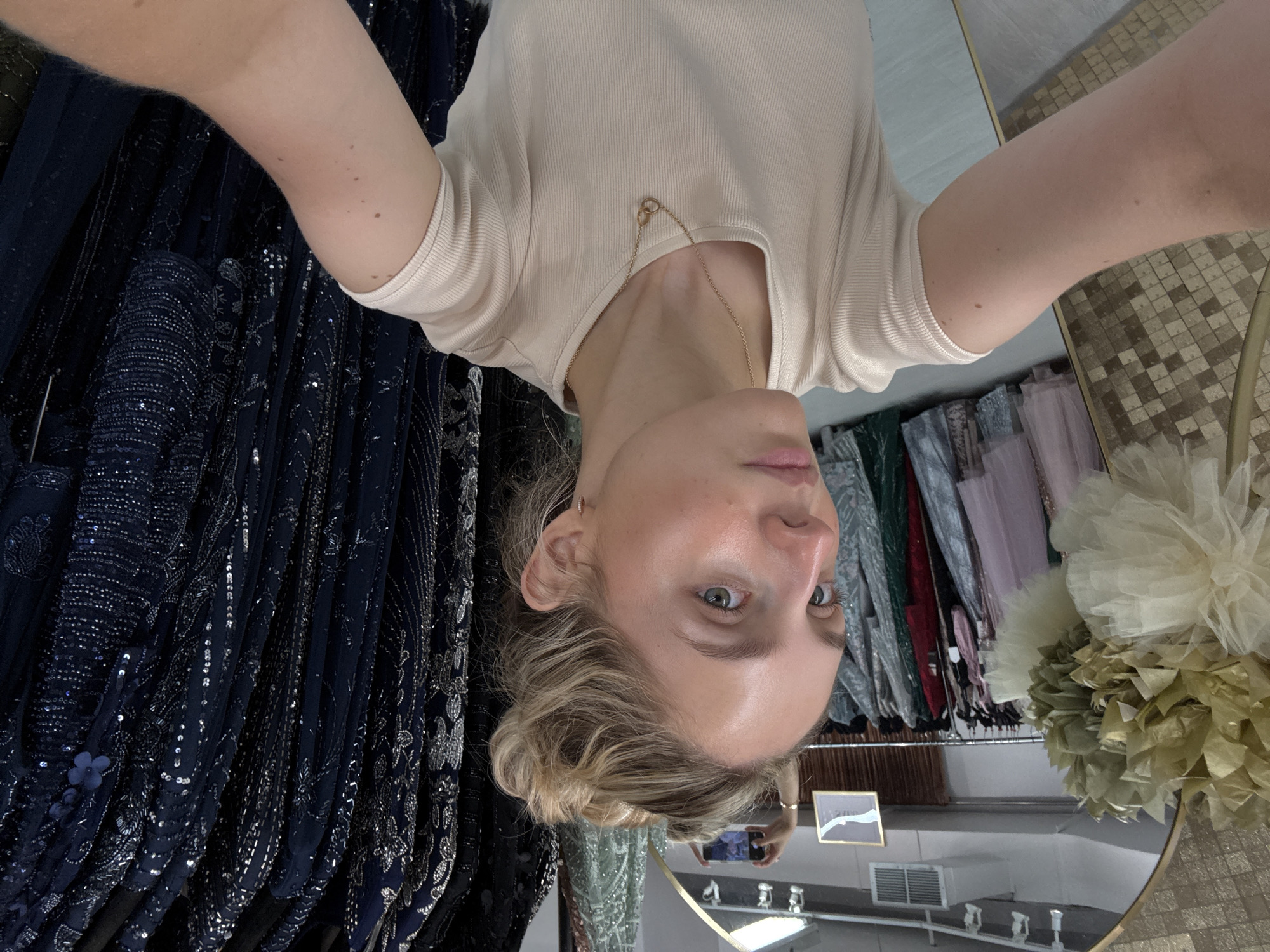 The image shows a young woman with blonde hair styled in a messy bun, taking a selfie in what appears to be a boutique or clothing store. She is wearing a cream-colored, ribbed t-shirt and a delicate gold necklace. The background reveals racks of formal dresses, predominantly in dark blue and embellished with sequins and intricate beadwork, suggesting a formal wear or evening gown store. To the left, there's a large decorative floral arrangement in gold and white, and a mirror reflecting the scene, including another phone held by someone in the background. The store environment is illuminated with overhead lighting. The photo appears to be taken in New York City.