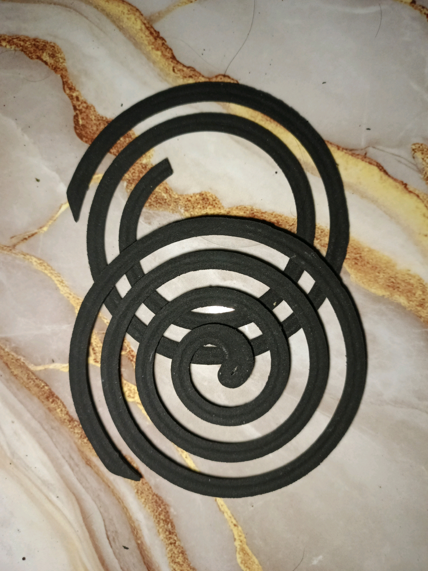 Two black mosquito coils are arranged in a stacked, overlapping spiral formation on a light-colored marble surface with brown and gold veining. The coils are positioned horizontally across the frame, with the top coil slightly offset to the left of the bottom coil. The background marble has a polished finish with natural, irregular patterns. No people or actions are depicted. The scene is static and focuses solely on the objects and their arrangement. The location is identified as Ketegan, Indonesia, though there are no visual cues within the image to support this geographical information.
