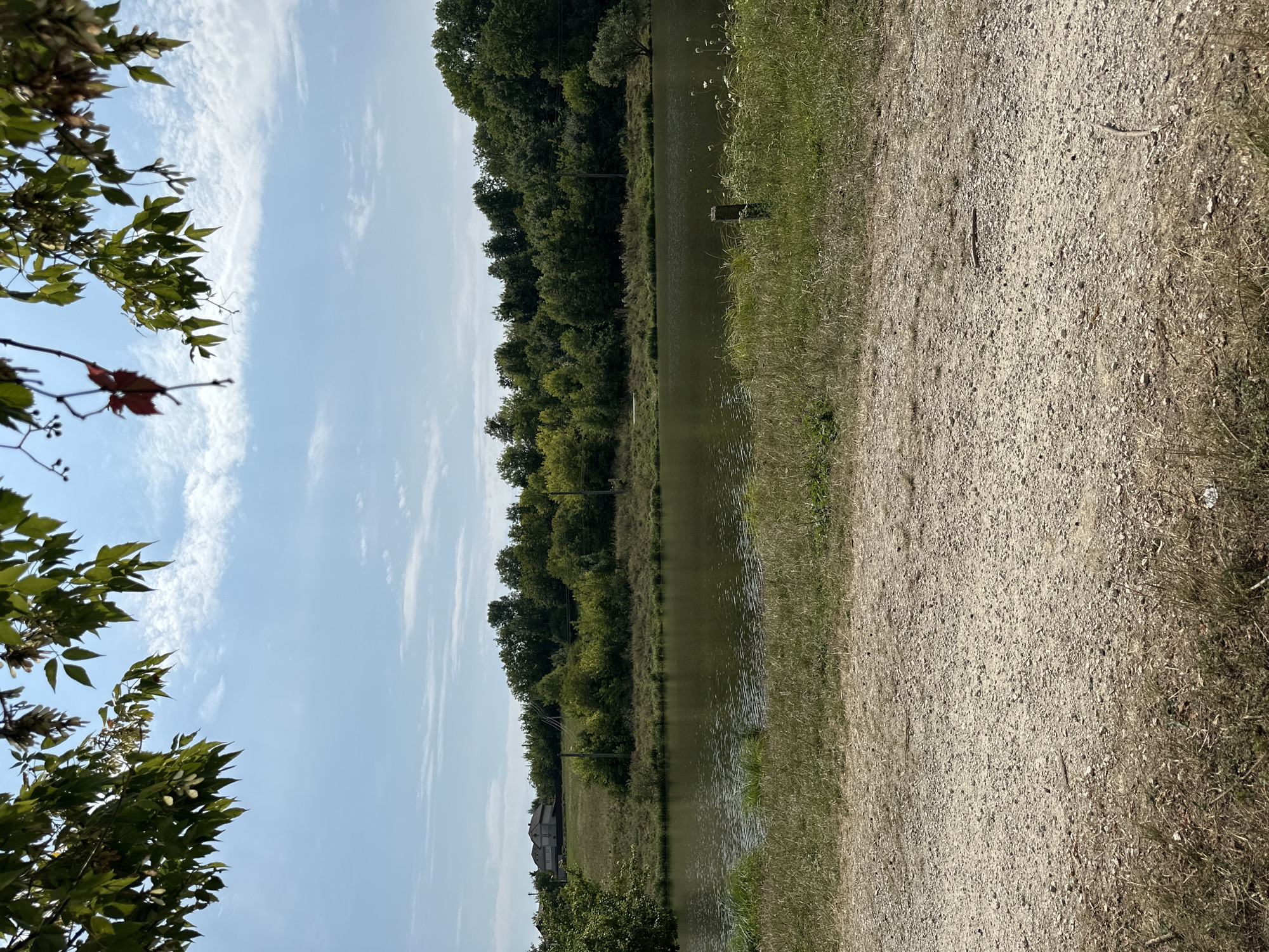 A serene landscape depicts a body of water with a lush green forest on the opposite bank. The foreground features a gravel path leading to the water's edge, with dry grass and scattered twigs. A wooden post stands near the water. In the distance, rolling hills are dotted with houses and trees under a partly cloudy sky. The image appears to be taken in Makiv, Ukraine.