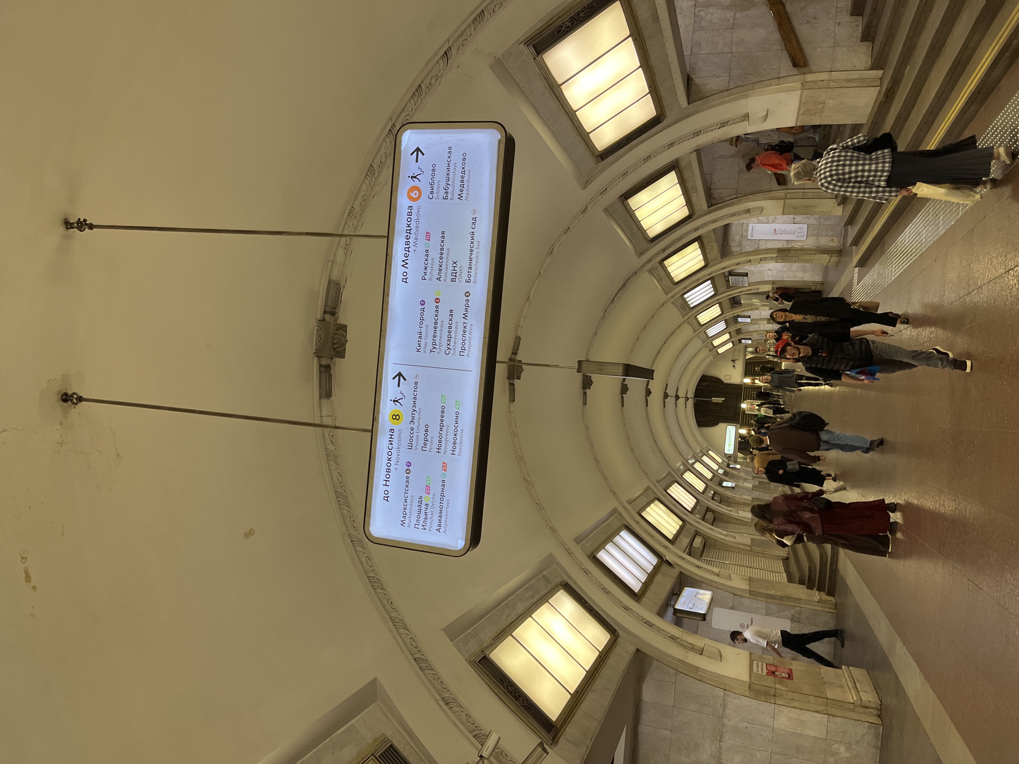 The image shows a view of a Moscow Metro station, specifically a section of the Sokolnicheskaya Line (Line 1) and the Kaluzhsko-Rizhskaya Line (Line 6). The signage indicates directions to Novokosino (Line 8) and Medvedkovo (Line 6). In the foreground, people are walking along the platform. The architecture of the station is characteristic of the Moscow Metro, with vaulted ceilings, arched passageways, and decorative elements. The lighting is provided by rectangular, illuminated panels set into the ceiling arches. The station appears to be clean and well-maintained. The scene is captured from a slightly elevated perspective, looking down the length of the platform.