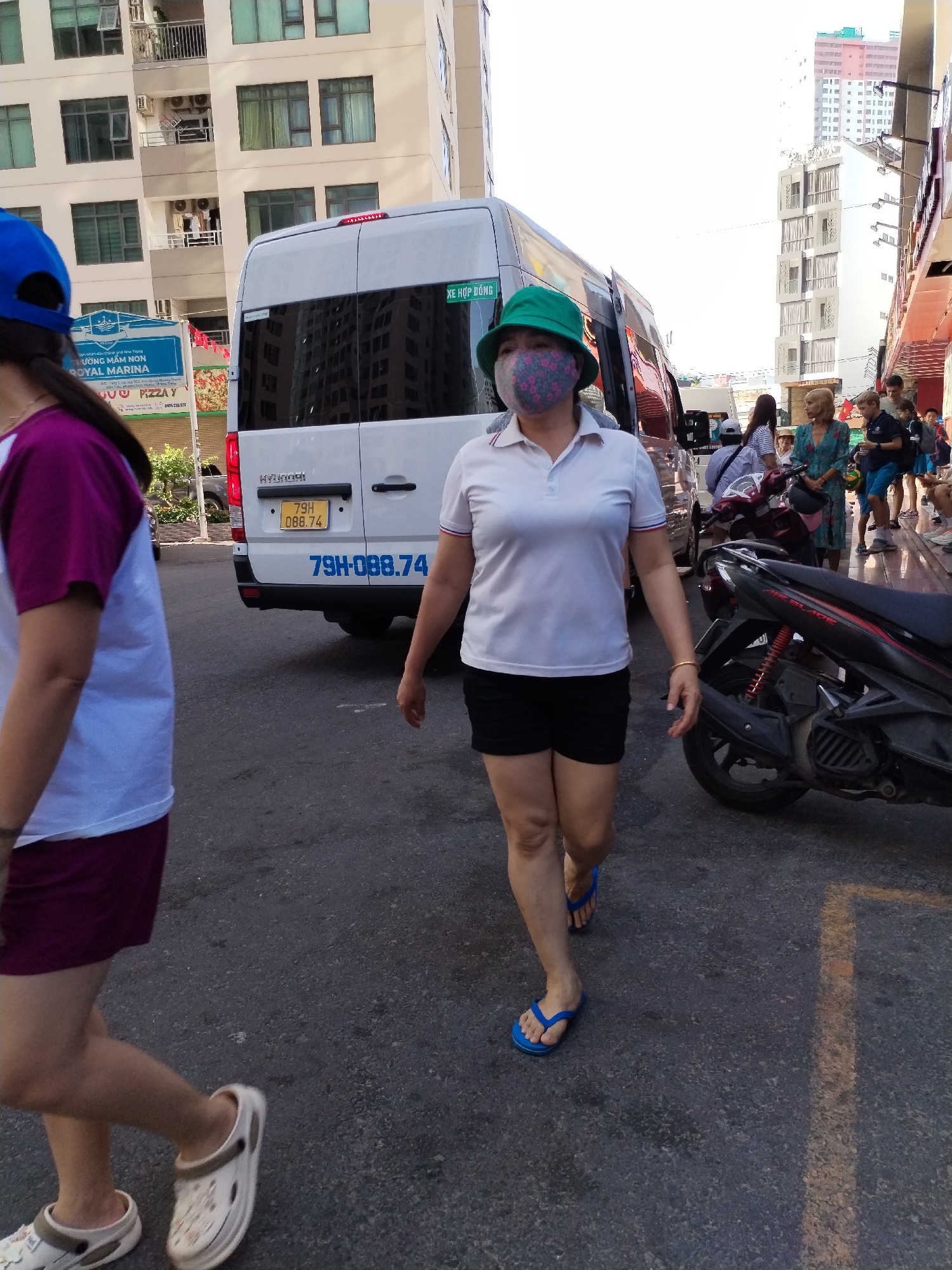 The image captures a street scene, possibly in the city of Bac Nha Trang Ward, Vietnam. A woman, wearing a green hat and a floral patterned mask, is walking towards the camera. She is dressed in a white polo shirt and black shorts, and she is wearing blue flip-flops. A second woman is walking on the left-hand side of the photo, her face is out of view. She is wearing a two-tone purple shirt and white Crocs. Behind the first woman is a white Hyundai van with license plate "79H-088.74", partially obscuring other pedestrians. A black motorcycle parked nearby. In the background is a building with several balconies. A pizza restaurant is on the left-hand side of the photo, with an advertising sign. The scene takes place on a paved street with a line of light running through the middle.