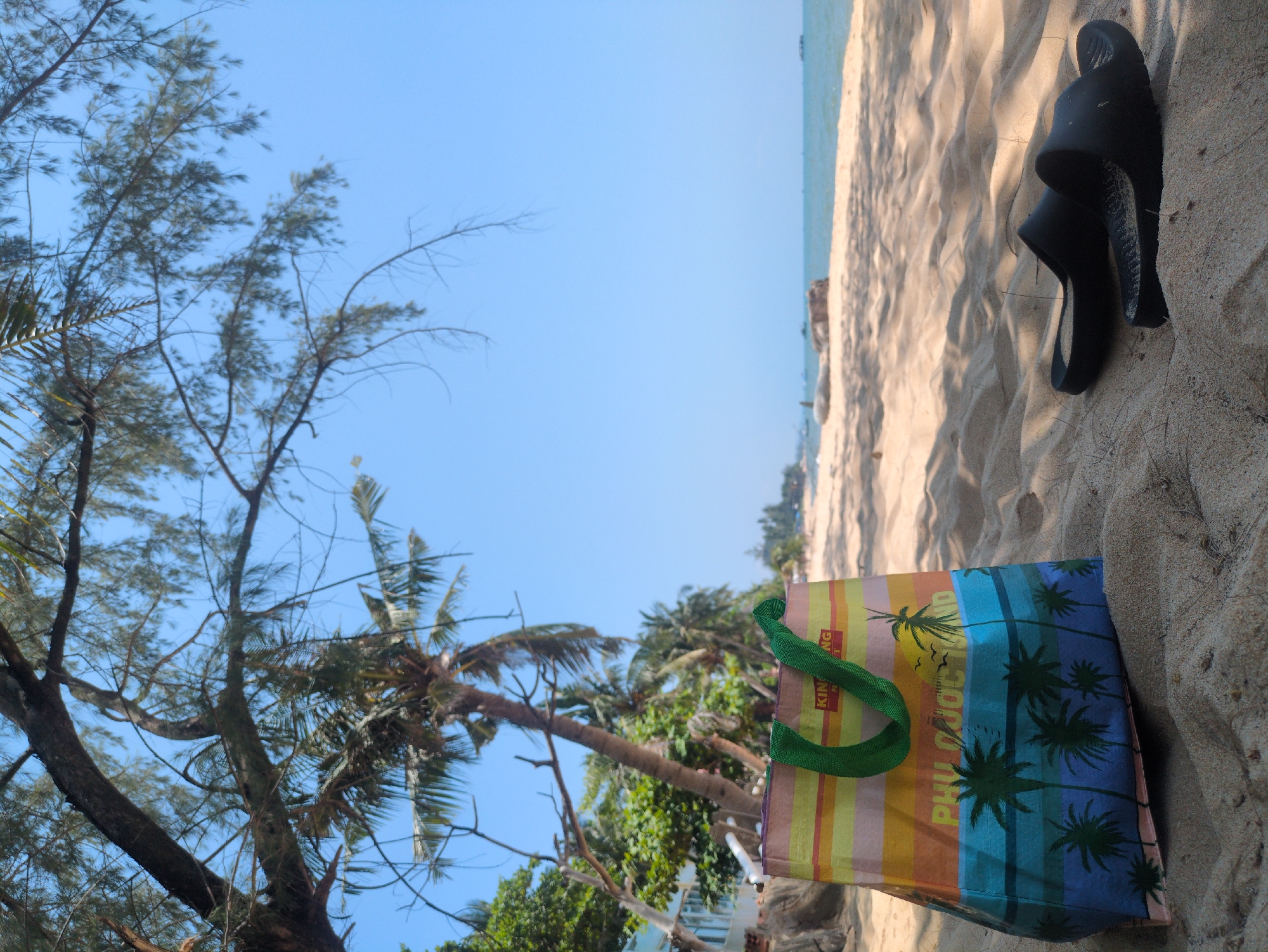 The image captures a scene on a beach in Phú Quốc, Vietnam. The sky is clear blue, contrasting with the sandy beach. Vegetation, including trees and palm trees, lines the shoreline. In the foreground, a colorful shopping bag rests on the sand, along with a pair of black sandals.
