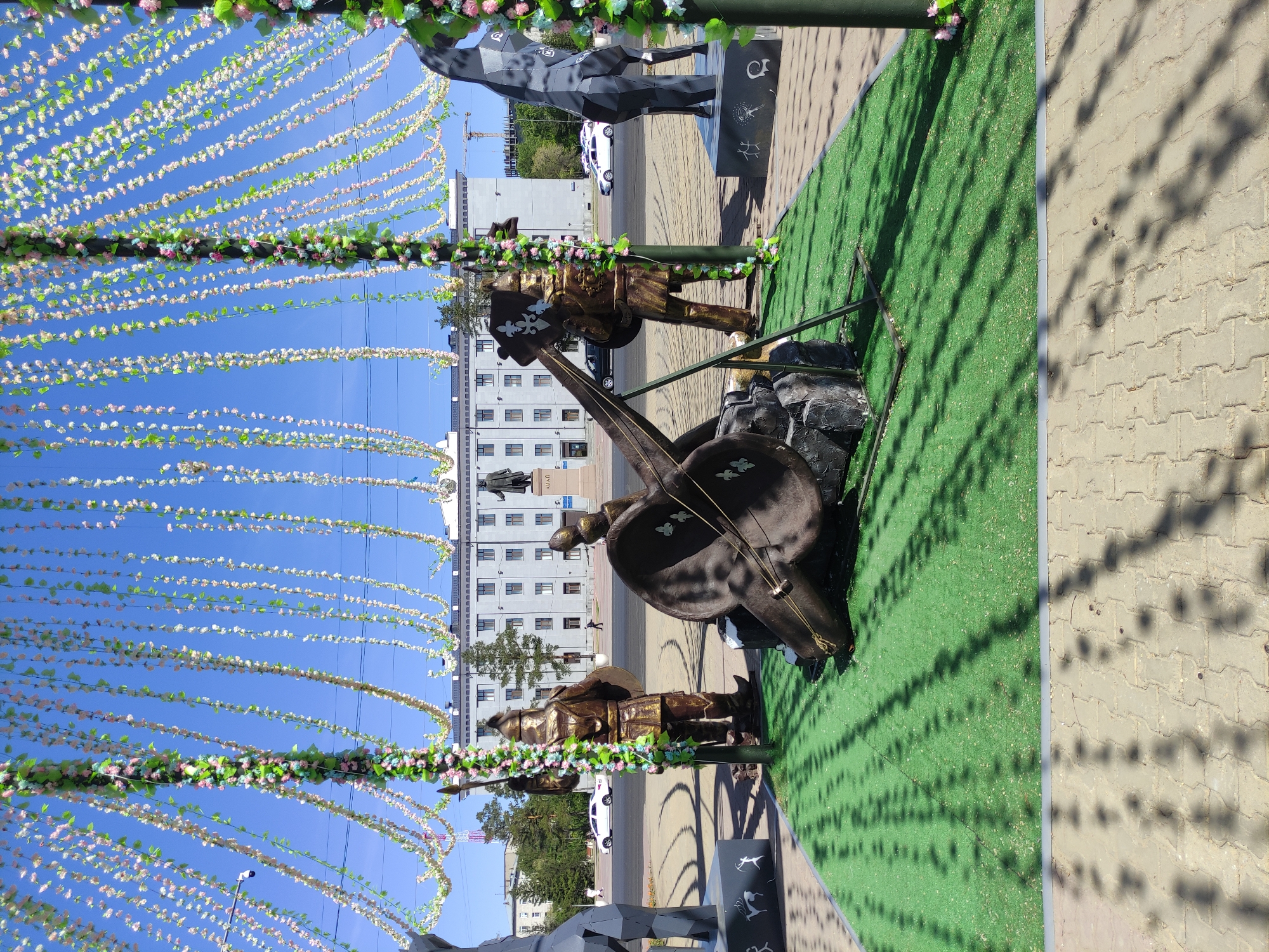 The image captures a street scene in Pavlodar, Kazakhstan, showcasing a bronze sculpture of musicians with a stringed instrument, possibly a dombra or a similar Central Asian lute, positioned on a patch of green artificial turf. The sculptures stand on display in what appears to be a public space. The architecture visible in the background includes a large, white building with multiple windows and a clock tower, likely a government or administrative building, indicative of a central city square. Above the sculptures, decorative garlands are strung across the street. The sky is a clear, bright blue. In the foreground, the edge of a paved walkway and shadow patterns on the ground are visible. A stylized geometric animal sculpture is also visible.