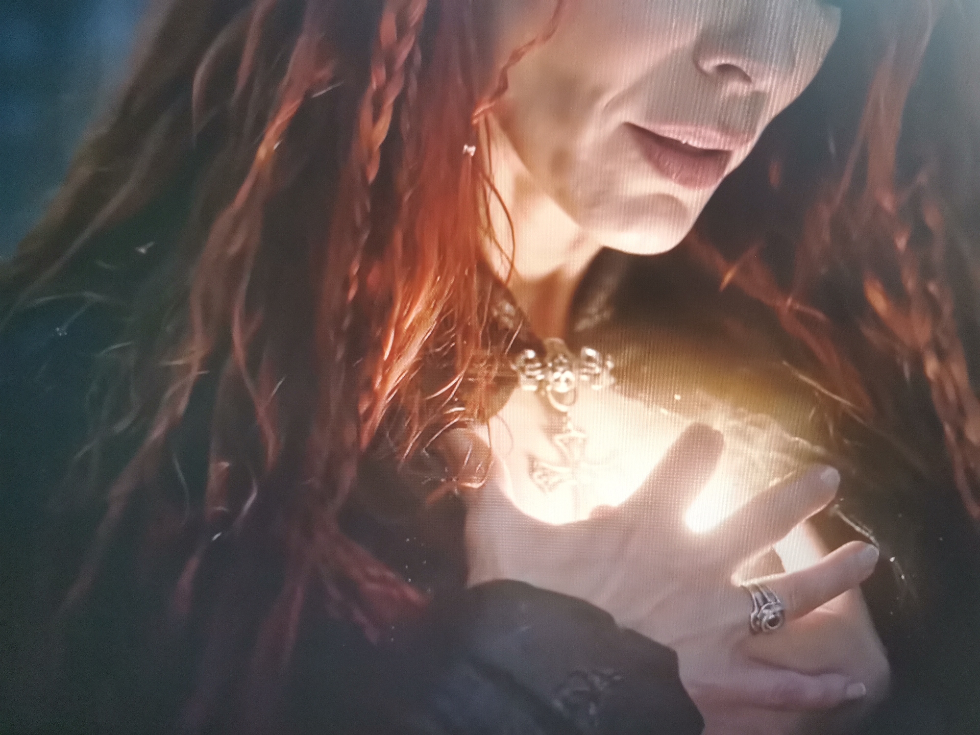 A close-up, medium shot captures a woman with long, reddish-brown, braided hair. She is wearing a dark garment and a detailed silver necklace featuring a skull and what appears to be a cross or ankh pendant. Her right hand is raised, cupped over her chest, where a bright, ethereal light emanates. A delicate silver ring with a dark stone is visible on her ring finger. The background is dark and out of focus, with hints of blue and green suggesting an outdoor or dimly lit environment. There is no discernible information within the image to indicate a specific location in Belaazyorsk, Belarus.