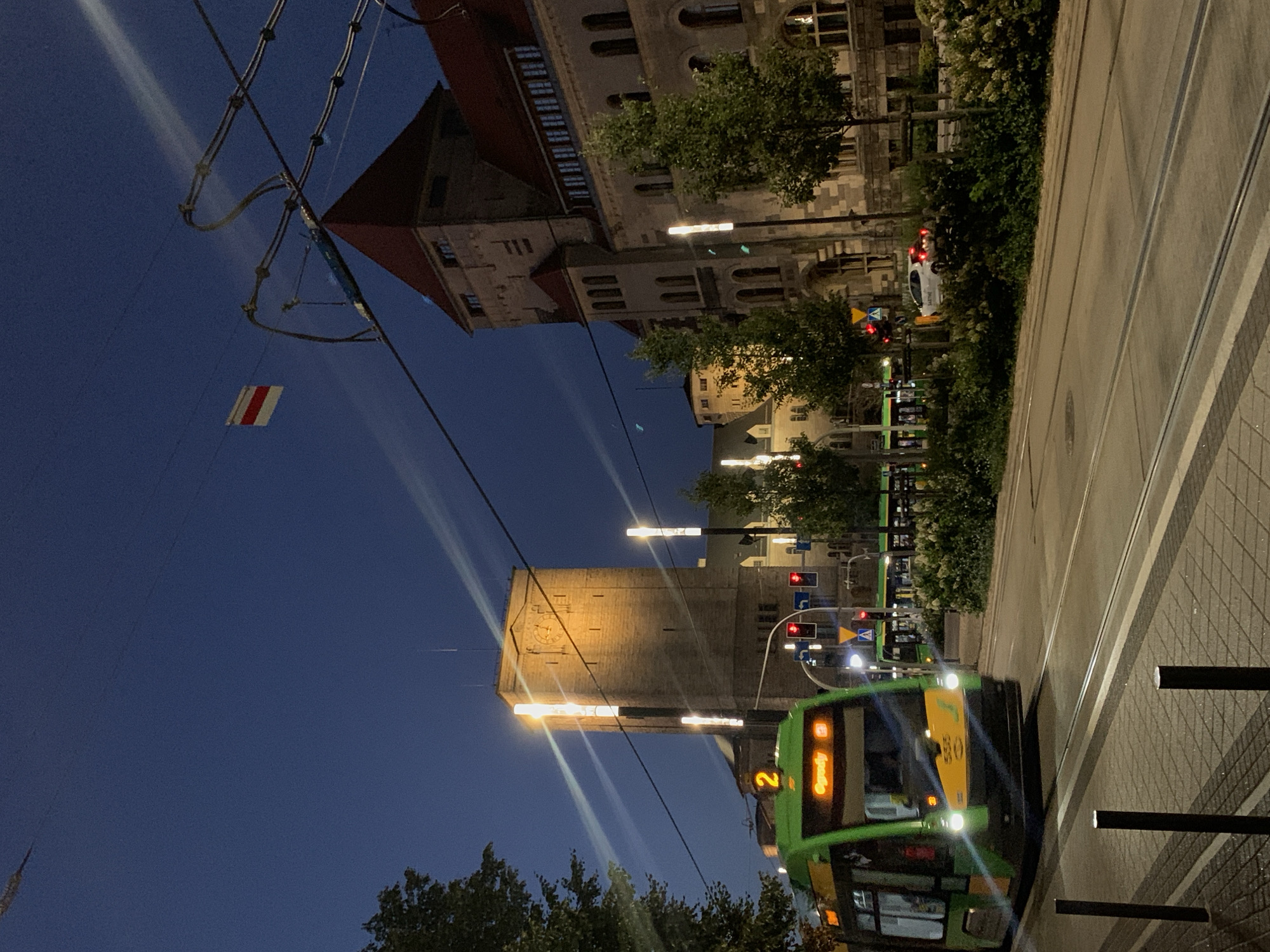 The image shows a night scene in Poznań, Poland. A green and yellow tram, numbered 2 and displaying "Ogrady" as its destination, is moving from left to right in the foreground. The tram has its headlights on and its interior is lit. In the background, a tall stone tower with a clock is visible, illuminated by lights. Adjacent to the tower is a building with a red-tiled roof and arched windows. The sky is dark blue, and there are overhead wires for the tram. A small white car is parked on the right side of the road, amidst greenery. The street is paved with cobblestones and has tram tracks. The overall atmosphere suggests an urban setting at dusk or night.