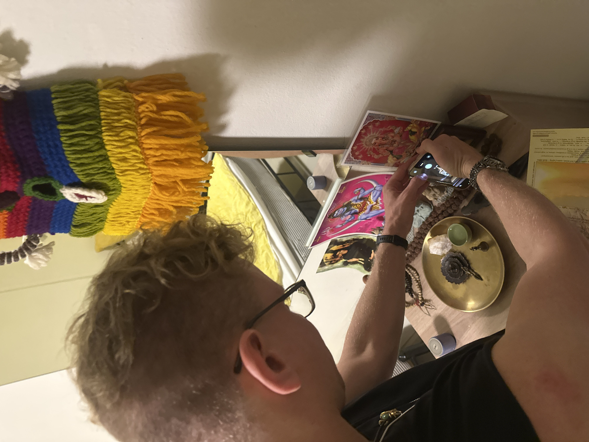 In an indoor setting, a person with curly blonde hair, wearing glasses and a black t-shirt, is leaning over a wooden surface, holding a smartphone to take a picture of items on the surface. The surface appears to be a shelf or table. On the surface are several items, including a golden plate with a dark, ornate object and a small green cup, a string of beads, and two framed pictures. One framed picture depicts a blue-skinned deity with multiple arms, and the other shows a deity with an elephant head. A colorful woven tapestry with fringe hangs on the wall behind the shelf. A mirror reflects a yellow-covered bed in the background. A red mark is visible on the person's left arm.
