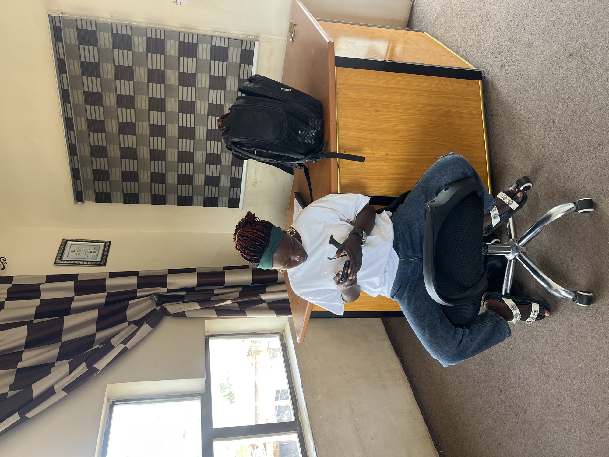A woman with braided red hair, wearing a green headband, a white t-shirt, and blue jeans, sits on a black rolling office chair at a wooden desk in a room in Jos, Nigeria. She is looking down and holding a black smartphone in her hands, with her thumbs actively engaged on the screen. A black backpack rests on the desk behind her. The room has a large window with brown and white checkered curtains on the left, and a patterned blind on the right. A framed certificate hangs on the wall above the curtains. The desk is made of light-colored wood with black trim. The floor is covered with a brown carpet.