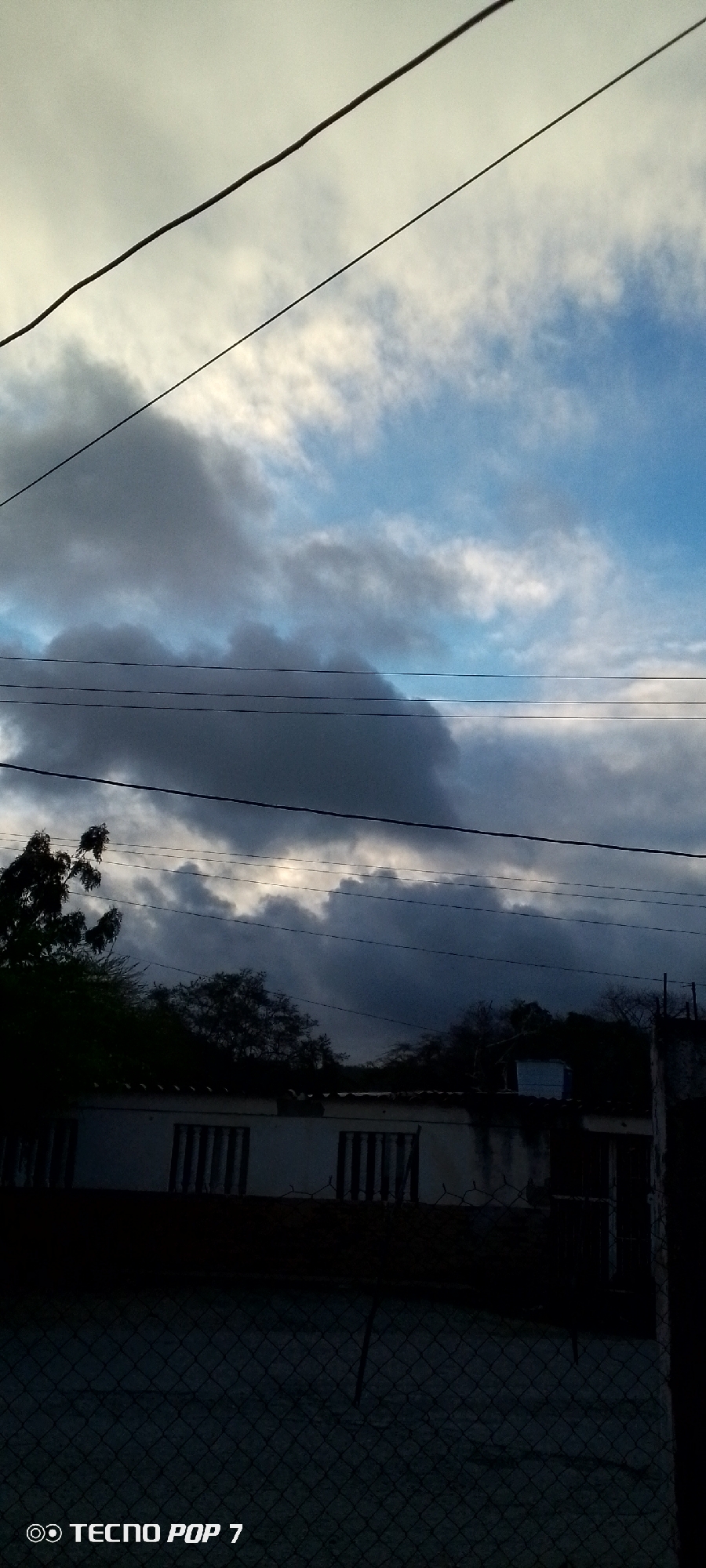 This image captures a sky filled with dramatic clouds, ranging from light and wispy to dark and heavy. Several power lines are visible, stretching horizontally across the frame, dividing the sky. Below the sky, a silhouetted row of buildings with barred windows is dimly visible. Trees and other vegetation obscure parts of the buildings and the horizon. In the foreground, a chain-link fence runs horizontally, and the bottom right corner of the image shows a portion of a pole. The bottom left corner of the image contains a watermark reading "TECNO POP 7". The scene appears to be an outdoor view in Altagracia, Venezuela, likely during a period of changing weather, given the cloud formations.