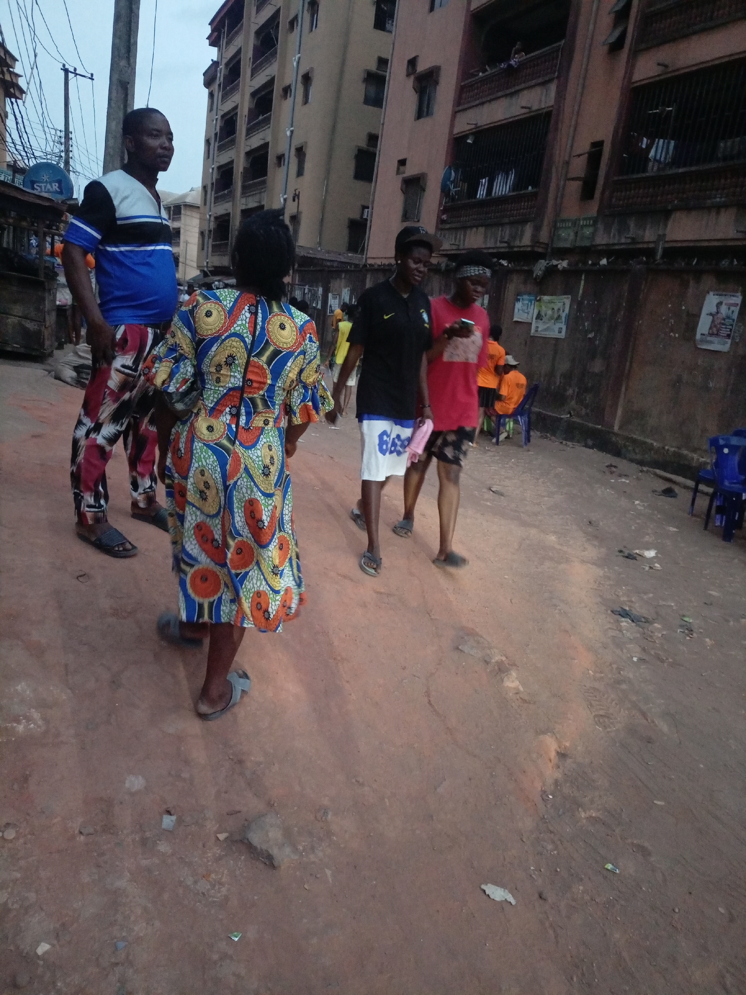 The scene is set in Onitsha, Nigeria, on a dirt road lined with buildings. A group of people are walking. A man in a blue and white shirt with patterned red pants stands to the left, talking to a woman facing away from the camera, wearing a brightly patterned blue, orange, and white dress. Two women walk to the right; one wears a black shirt with white shorts, the other wears a red shirt and a bandana. A group of people in orange shirts are sitting on blue chairs near a wall covered in posters. The building in the background is tall and appears to have barred windows. The sky is overcast.