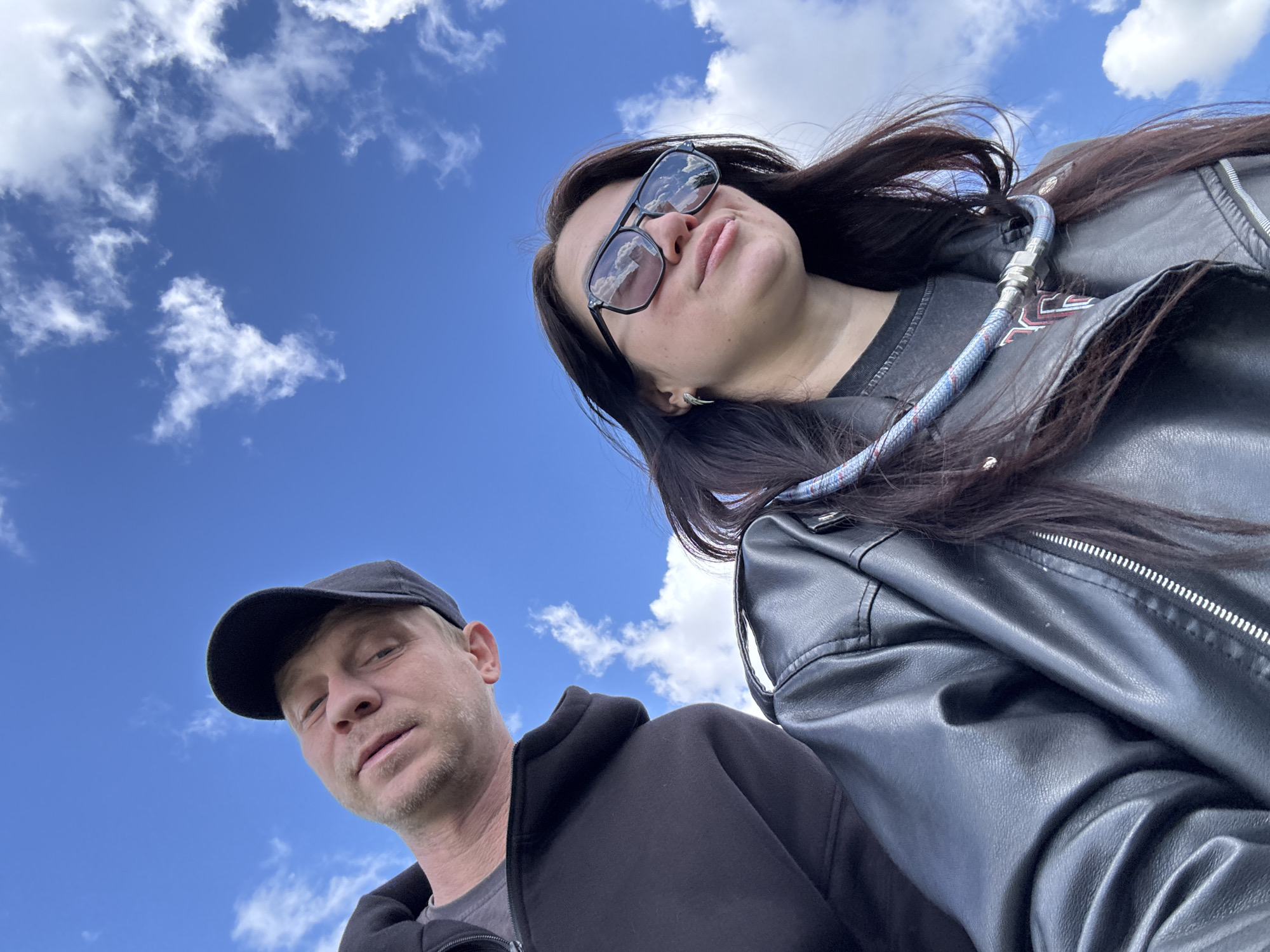 This is a low-angle outdoor portrait featuring two individuals against a bright blue sky with scattered white clouds.
In the upper right portion of the frame, a woman is visible from the chest up. She has long, dark hair flowing over her shoulders and is wearing dark sunglasses that reflect the clouds and blue sky. She is dressed in a black leather jacket over a dark top. A blue and white braided hose or cable with a metallic connector is draped across her shoulder and chest. Her expression is neutral.
In the lower left, a man is partially visible, also from the chest up. He is wearing a black baseball cap and a dark hooded jacket over a gray t-shirt. He has light stubble on his face and a neutral expression, looking directly at the camera.
The composition places both subjects looking down towards the camera, suggesting a selfie taken from a low vantage point, possibly held by one of them. The background is dominated by a clear, vibrant blue sky dotted with fluffy white clouds, indicating daytime and good weather. There are no discernible location cues or visible text within the image.