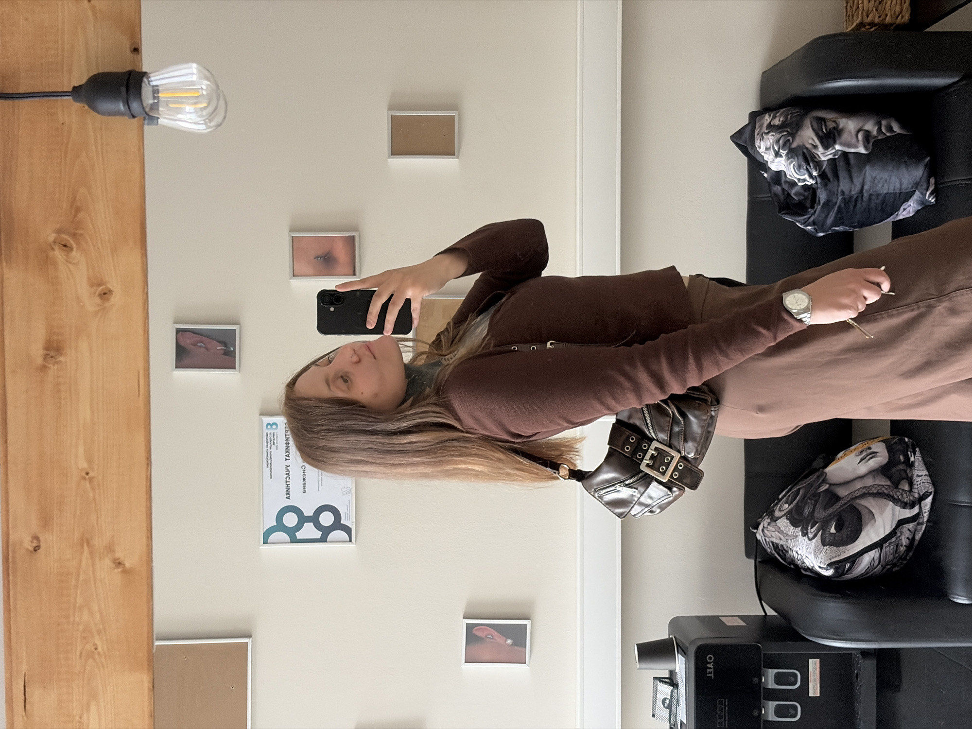 The image shows a young woman with long brown hair taking a mirror selfie indoors. She is wearing a brown long-sleeved shirt and a brown skirt, and she has a brown purse slung over her shoulder. She is holding a black smartphone in her right hand. In the background, there are framed pictures on the wall and a wooden beam across the top. A black leather sofa is visible in the lower part of the image, with two decorative pillows on it, one with a black and white graphic pattern and another with a sculpture of a human head. To the left of the sofa is a water cooler. The room appears to be a waiting area or lounge. The location is not identifiable as Chaikovsky, Russia, based on the visual information.