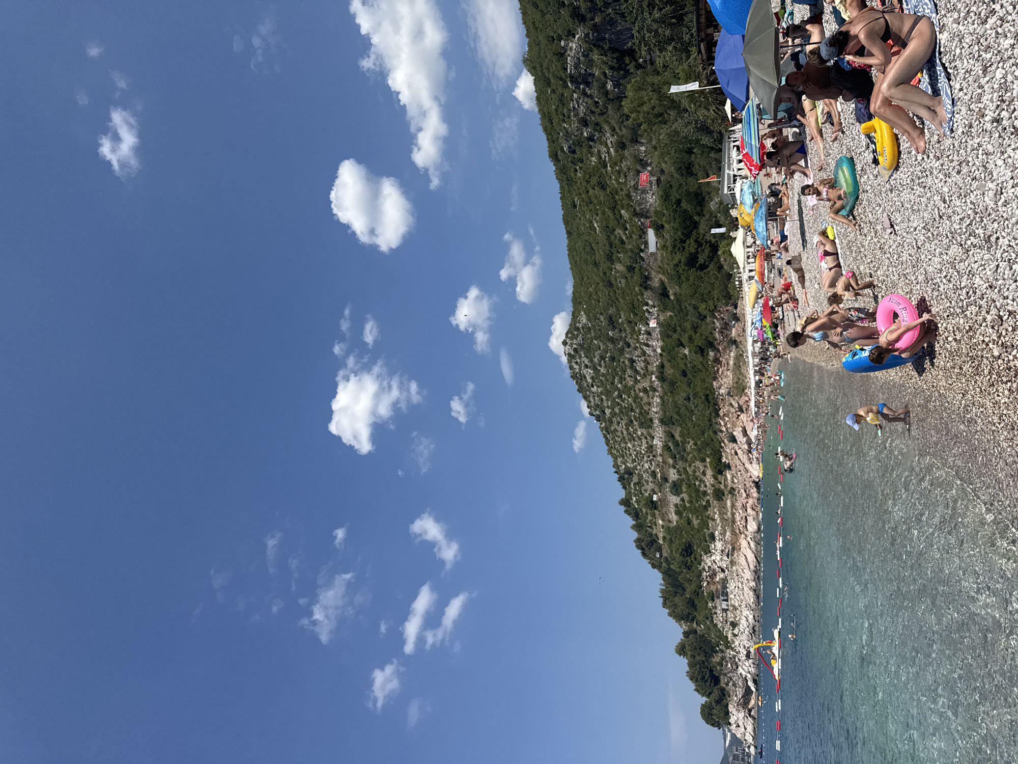 The image depicts a crowded pebble beach on a sunny day, likely in the summer. A steep, forested mountain slopes down to the edge of the beach, with a clear blue sky dotted with white clouds above. The sea is calm and translucent, with people swimming and wading. Beach umbrellas provide shade for sunbathers lounging on towels and inflatable rings. Several people are visible in the water, including children with inflatable toys. The scene suggests a leisurely day at the beach, with a vibrant atmosphere. There are some signs and structures visible on the beach, including what appears to be a small inflatable water park. Some text is visible on the pink inflatable ring in the foreground, which reads "Swim Ring". The time of day appears to be midday, given the strong sunlight and the shadows cast by the umbrellas. The overall impression is one of a popular tourist destination offering a classic beach experience.